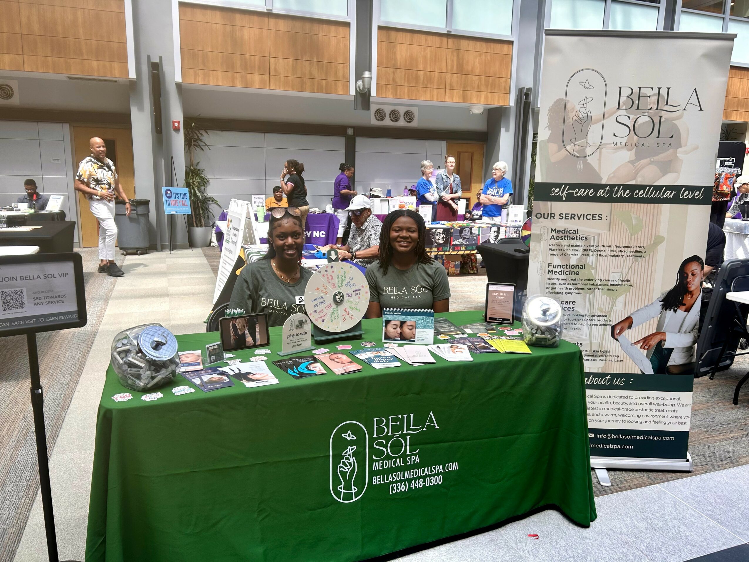 Two people sit behind a table with a green tablecloth displaying information about Bela Sol, in a busy indoor space with people and banners.