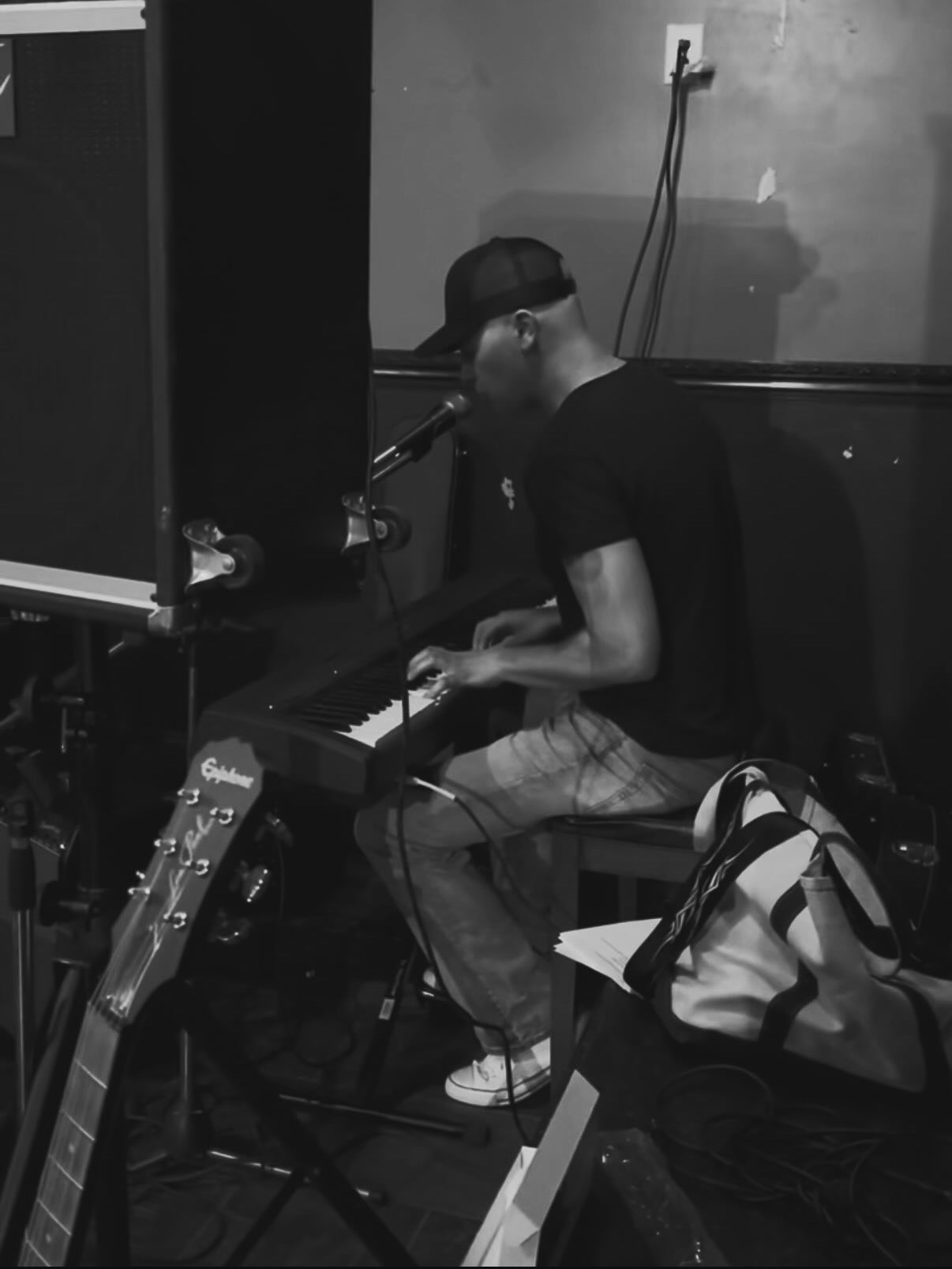 Person playing keyboard in a room with musical equipment, wearing a cap and glasses, with a backpack nearby.