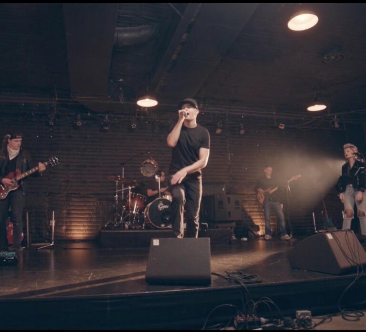 Band performing on stage with five members, one singing into a microphone, in a dimly lit venue.
