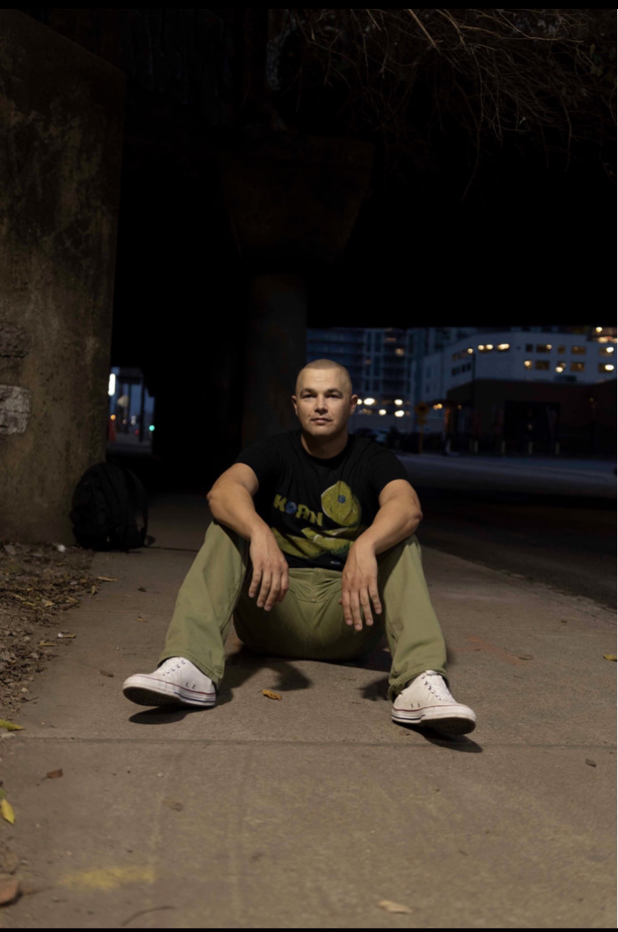 Person sitting on sidewalk at night, with buildings and streetlights in background.