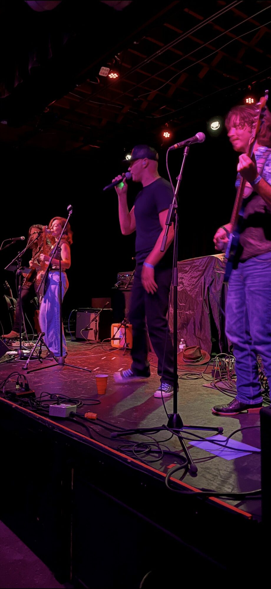 Group of young people performing on stage with microphones and musical instruments, colorful stage lighting, and a dark background.