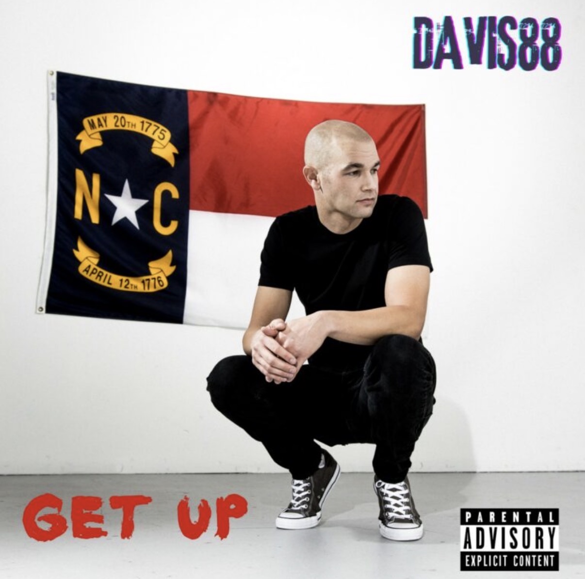 Person crouching in front of a flag with a red, black, and white design, with text and a star symbol, on a white wall.