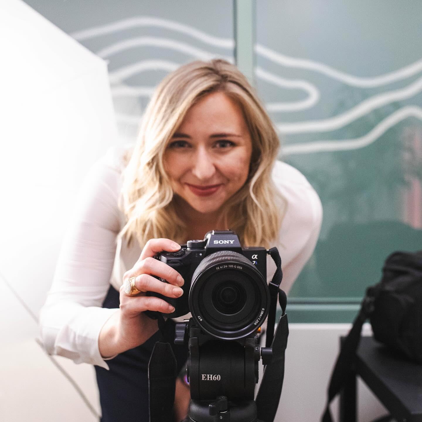Woman with blonde hair taking a selfie with a camera, smiling, in a room with abstract wall art.