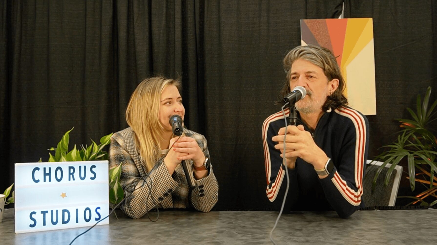 Two people sit at a table with microphones, smiling and talking, with a sign reading 'CHORUS STUDIOS' and plants in the background.