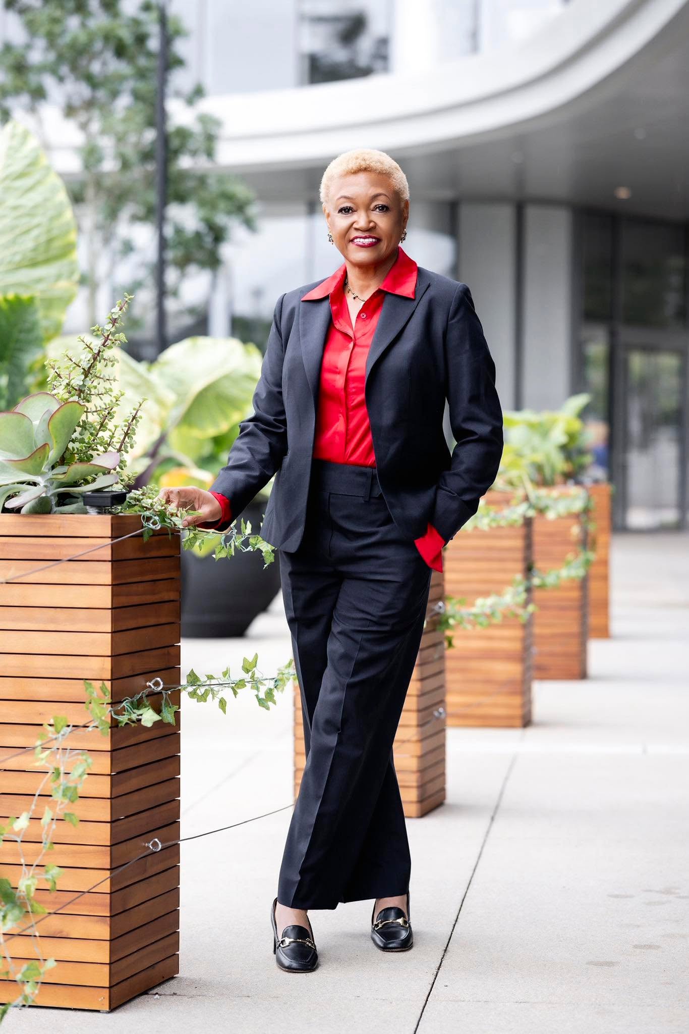 Smiling woman in a navy suit and red shirt standing outdoors near planters with greenery, modern building in background.