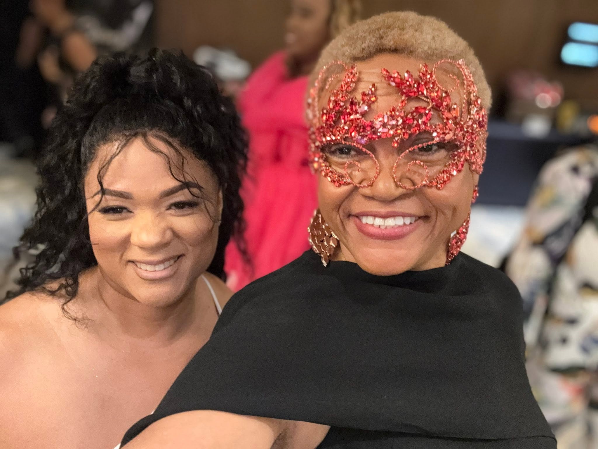 Two women smiling at an indoor event, one wearing glasses with red embellishments and a black top, the other with curly hair.