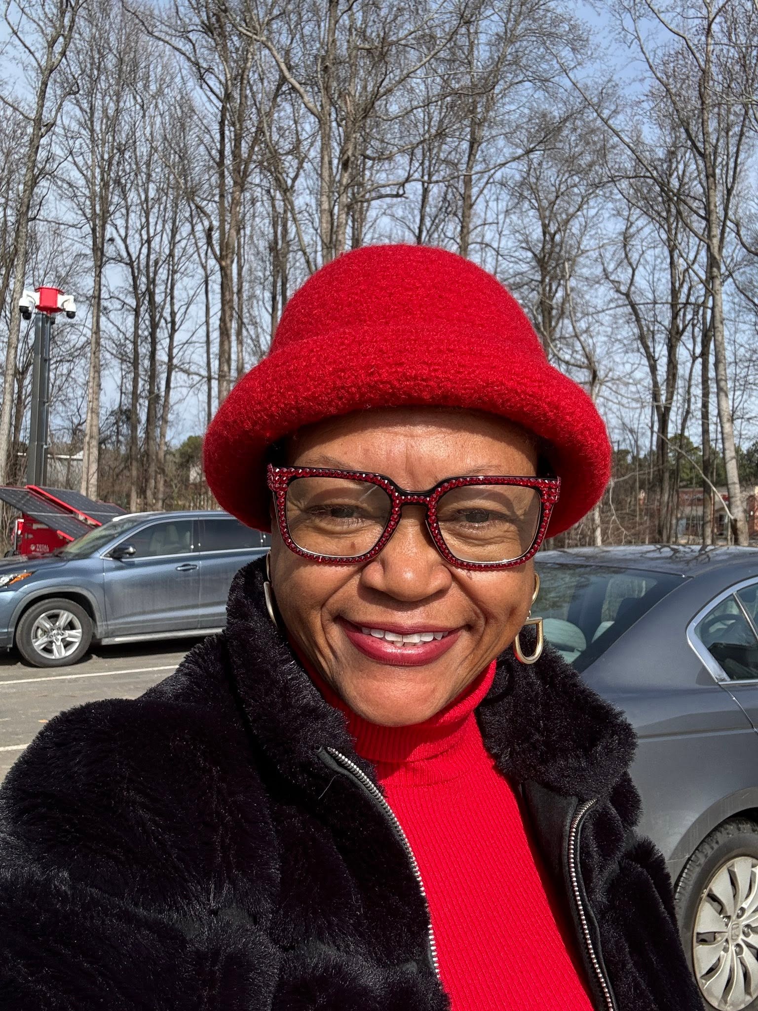 Woman outdoors wearing red hat and glasses, smiling, with parked cars and leafless trees in background.