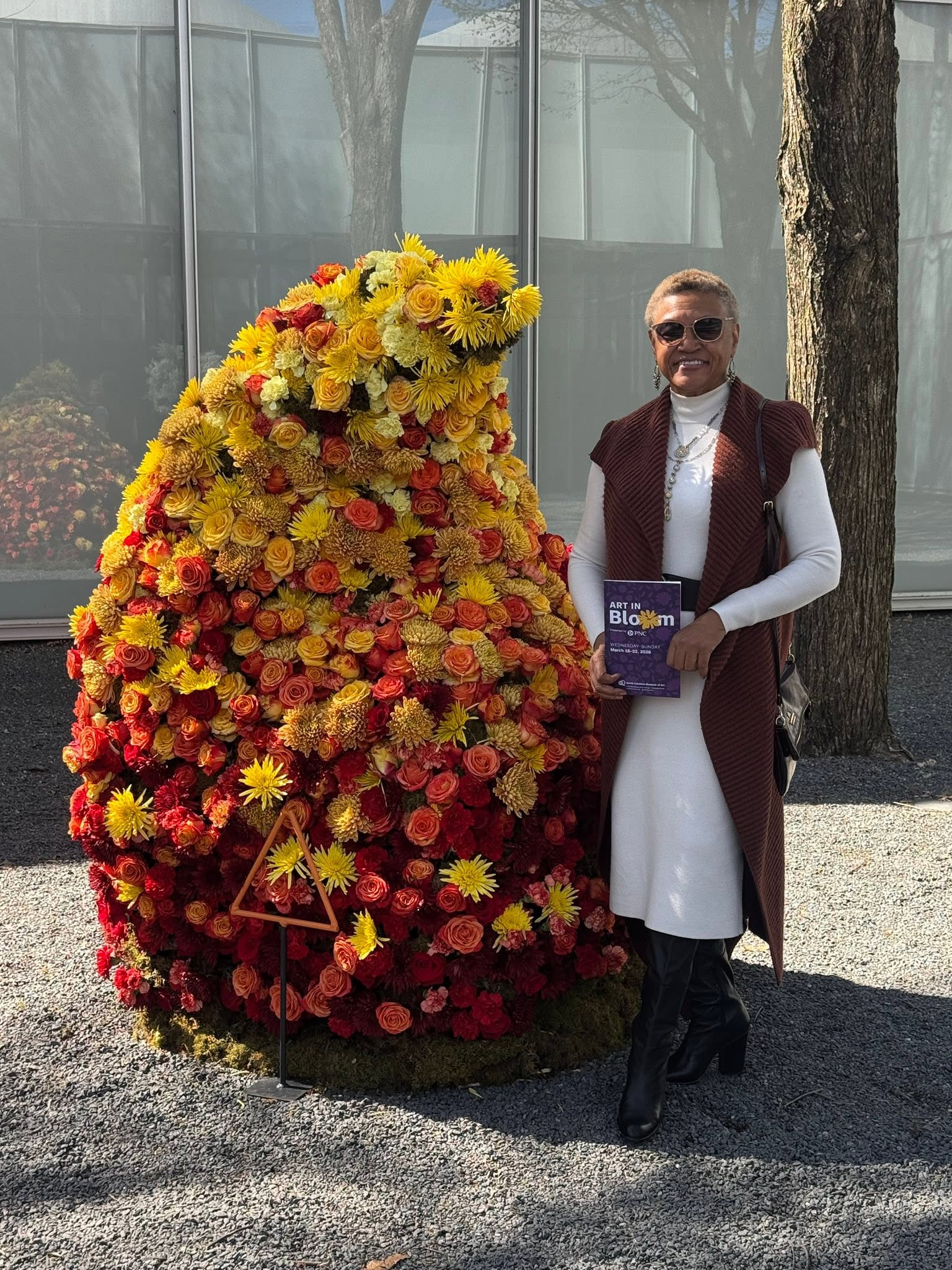 Person standing next to a large flower arrangement shaped like a rabbit, holding a book, outdoors with trees and a glass building.