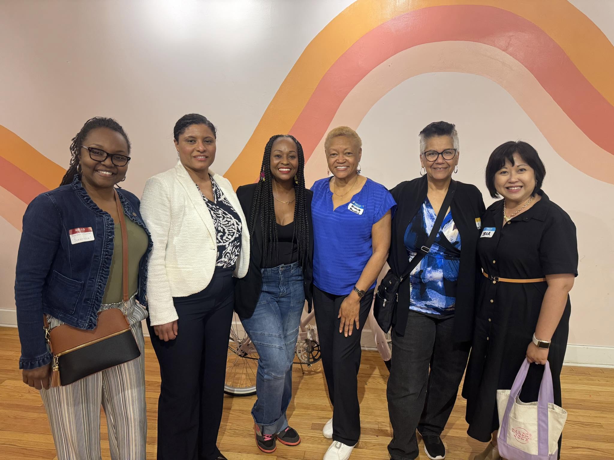 Group of seven women standing indoors in front of a colorful rainbow mural, smiling, with wooden flooring visible.