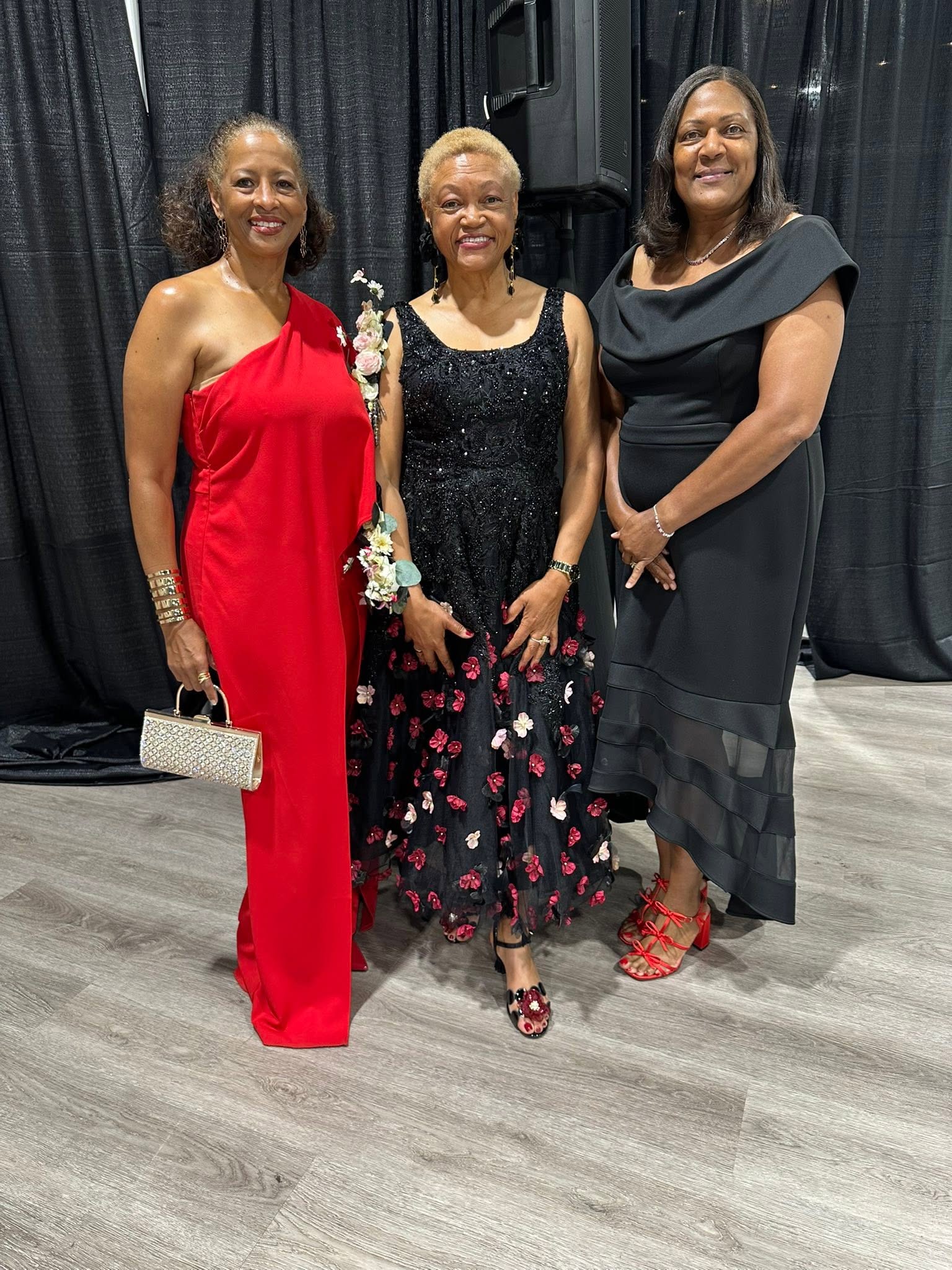 Three women in formal dresses standing together indoors, smiling, with dark curtains in the background.