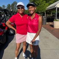 Two women in golf attire standing outdoors, smiling, with golf carts and trees in background.