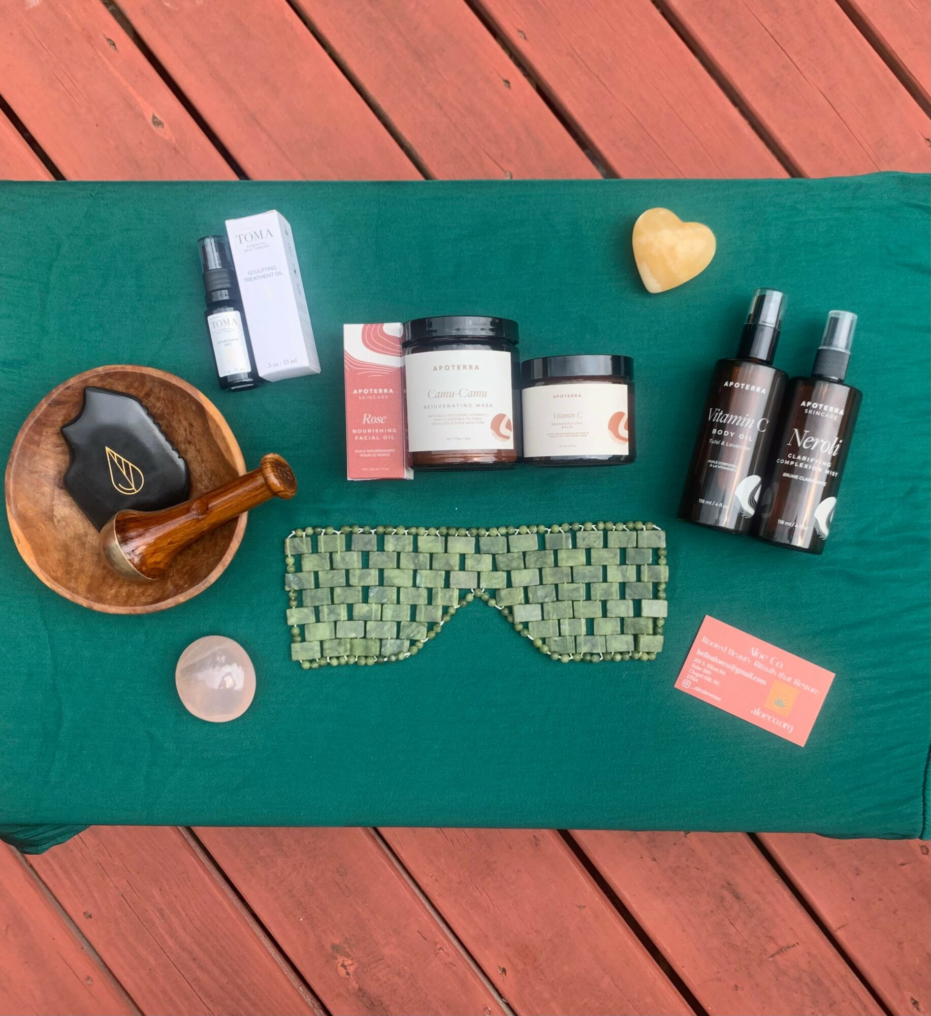 Skincare and wellness products arranged on a green cloth, with a wooden bowl, heart-shaped object, and a patterned eye mask.