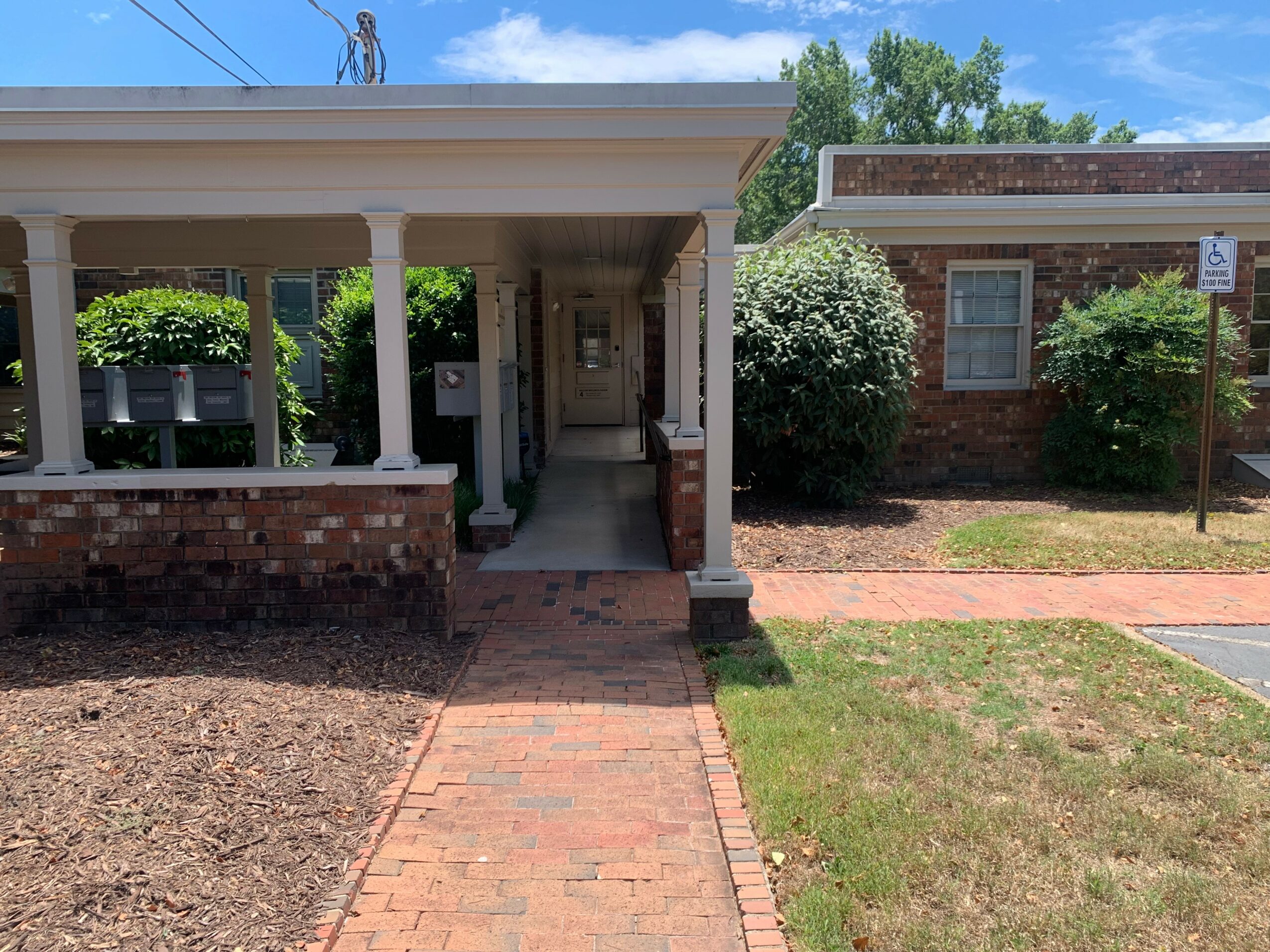 Front view of a house with a porch, brick walkway, bushes, and a brick building in the background.