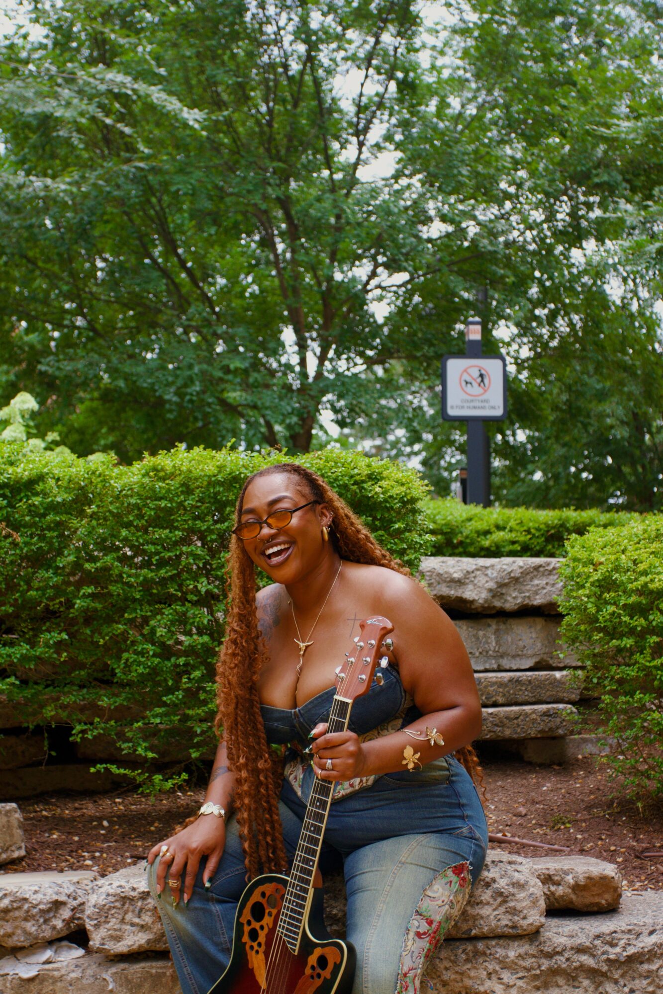 Woman with long braided hair and glasses sitting on stone steps holding a guitar outdoors, surrounded by greenery.