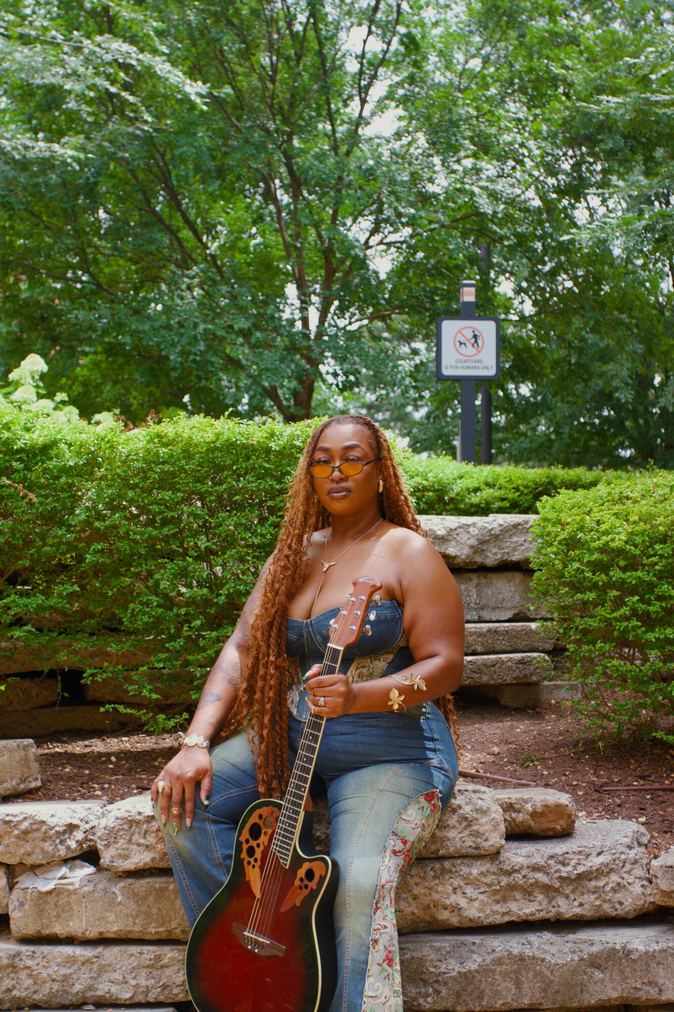 Woman with long hair and glasses holding a guitar, sitting on stone steps in a park with green trees and bushes.