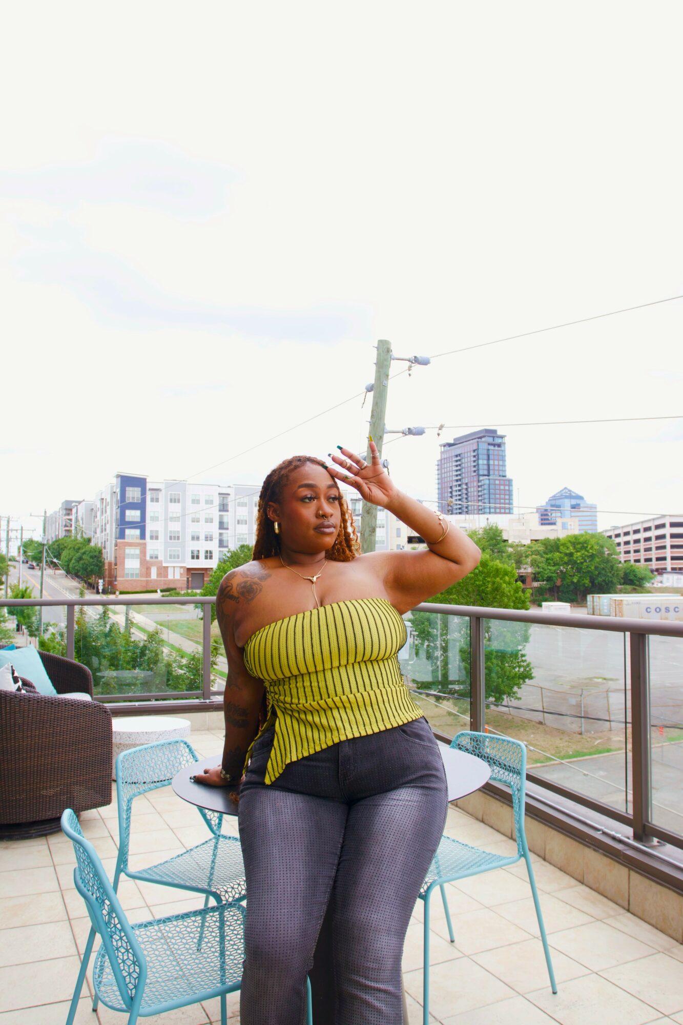 Woman with long hair in a yellow strapless top and dark pants sitting on a balcony with city buildings in the background.
