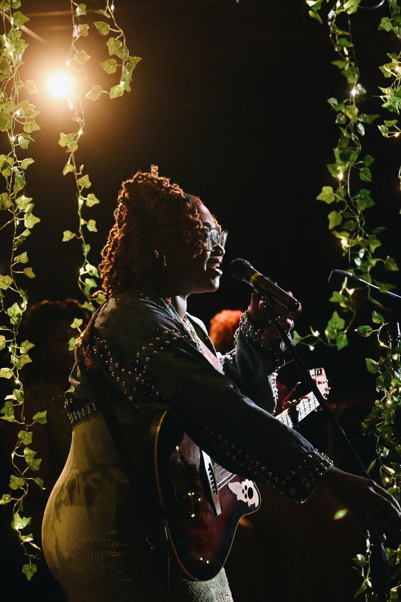 Person with curly hair singing into a microphone on stage, surrounded by hanging plants, with sunlight shining from the top left.