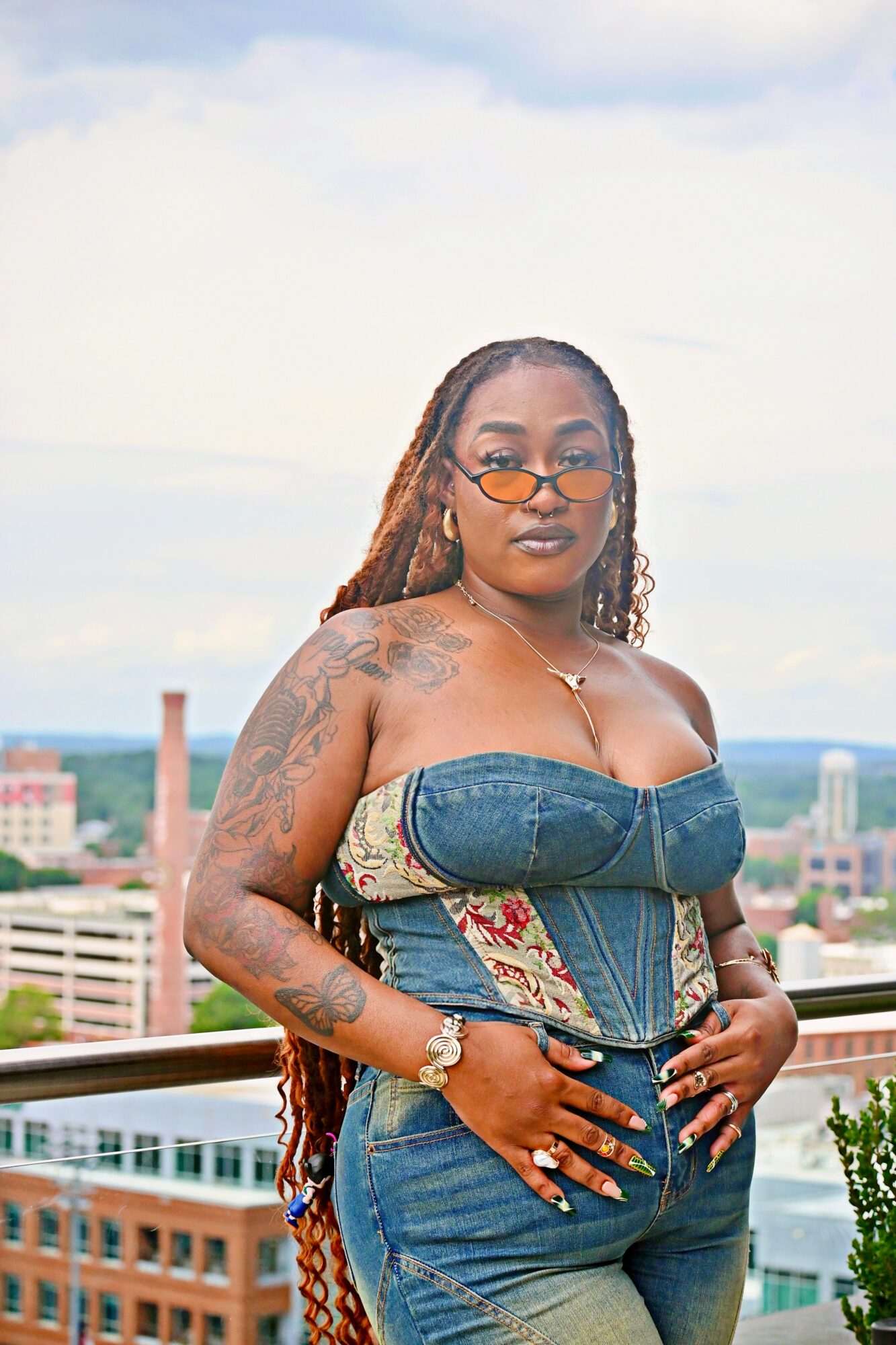 Woman with tattoos and glasses standing outdoors on a balcony with cityscape background, wearing a strapless denim top and jeans.