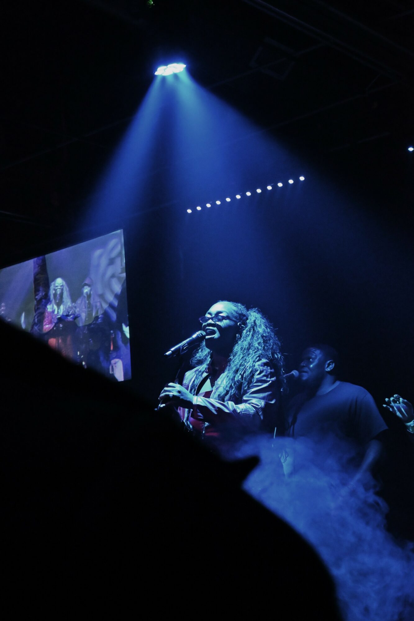 A singer with curly hair performs on stage under blue lighting, with a large screen and fog effects in the background.