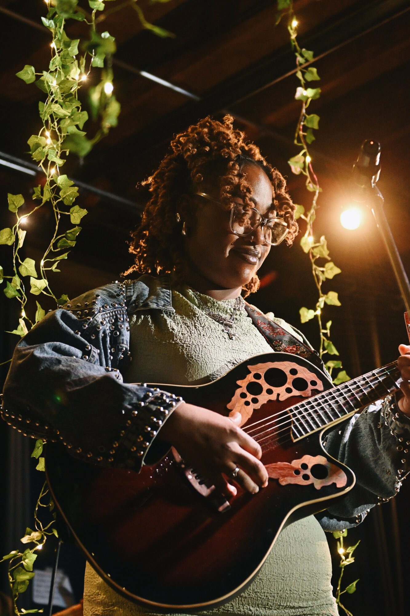 Person with curly hair playing an acoustic guitar, surrounded by hanging green plants, with warm lighting in the background.