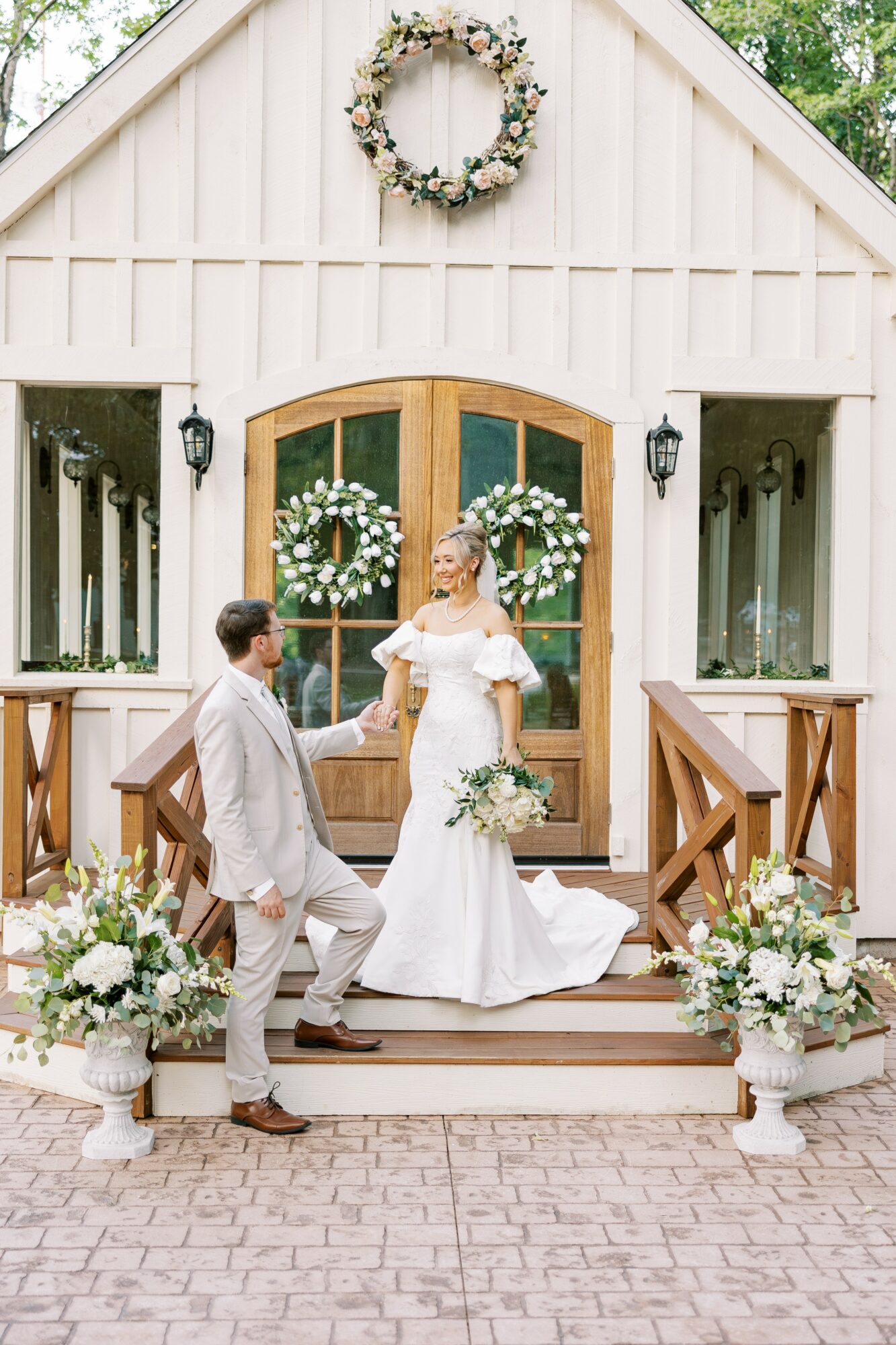 Bride and groom stand on steps outside a white building with floral wreaths on door, holding hands, surrounded by flower arrangements.