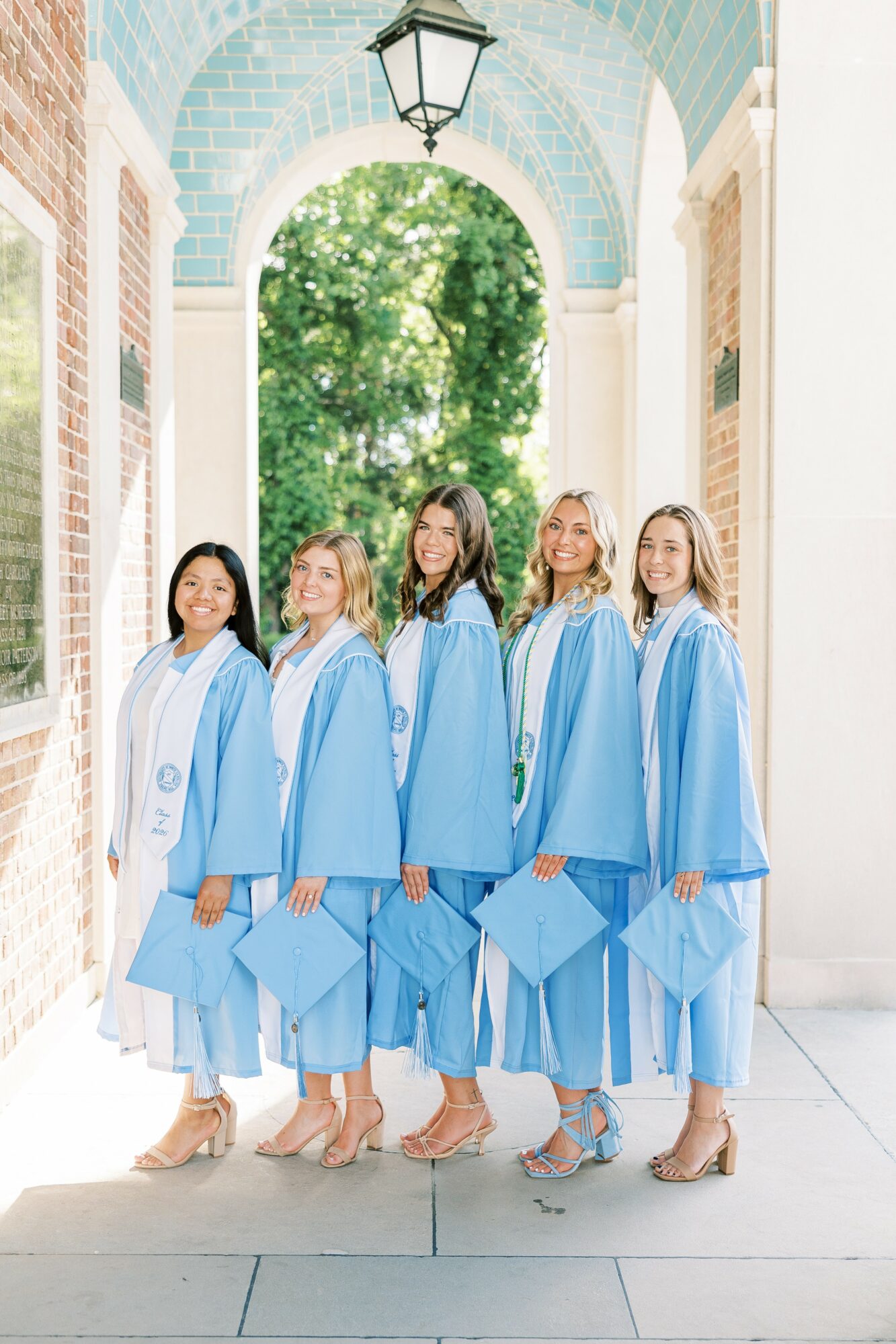 Five women in blue graduation gowns and caps stand outdoors under an archway, smiling, with trees in the background.