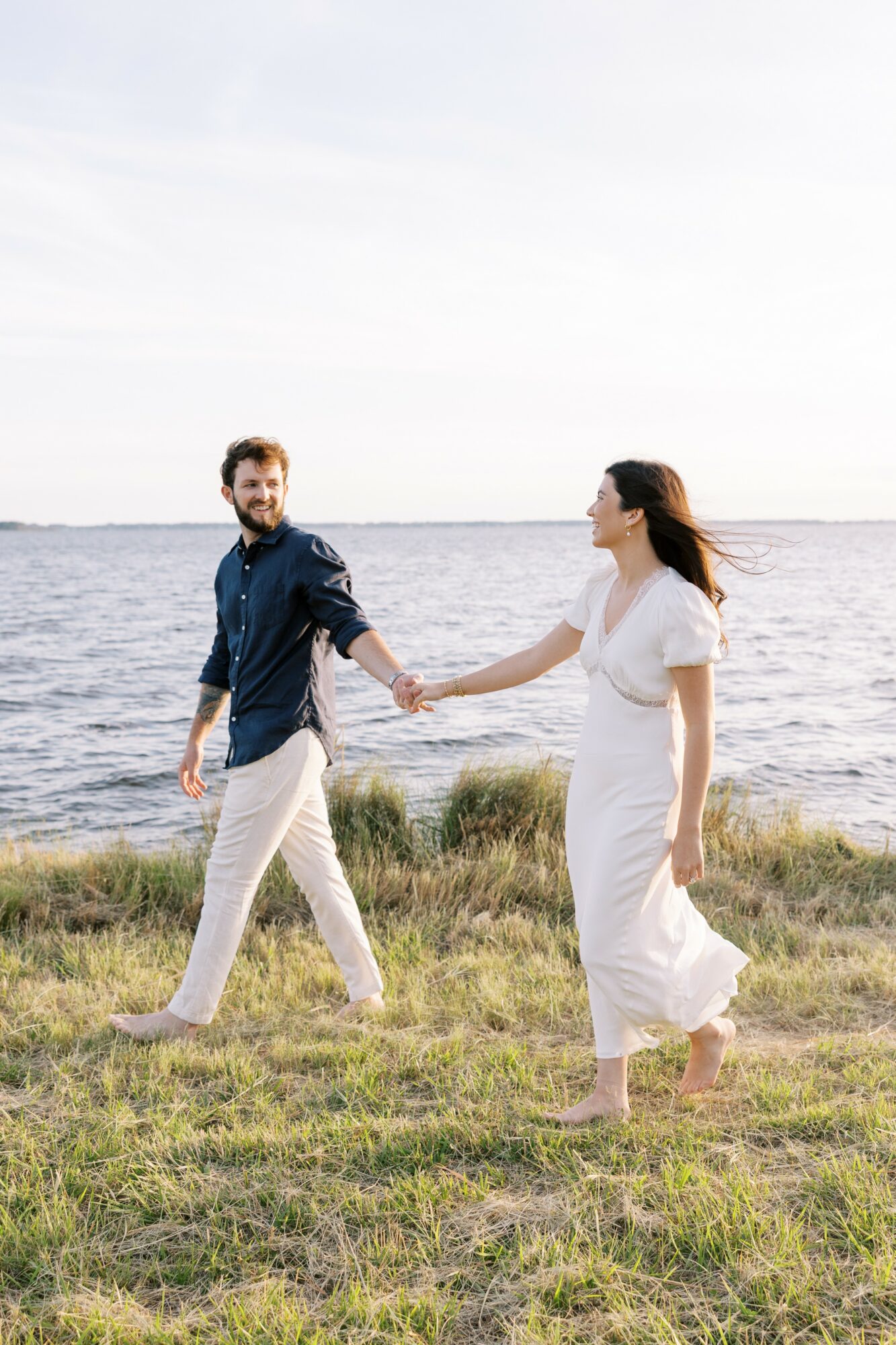 A couple holding hands walking on grass near water, with a cloudy sky in the background.