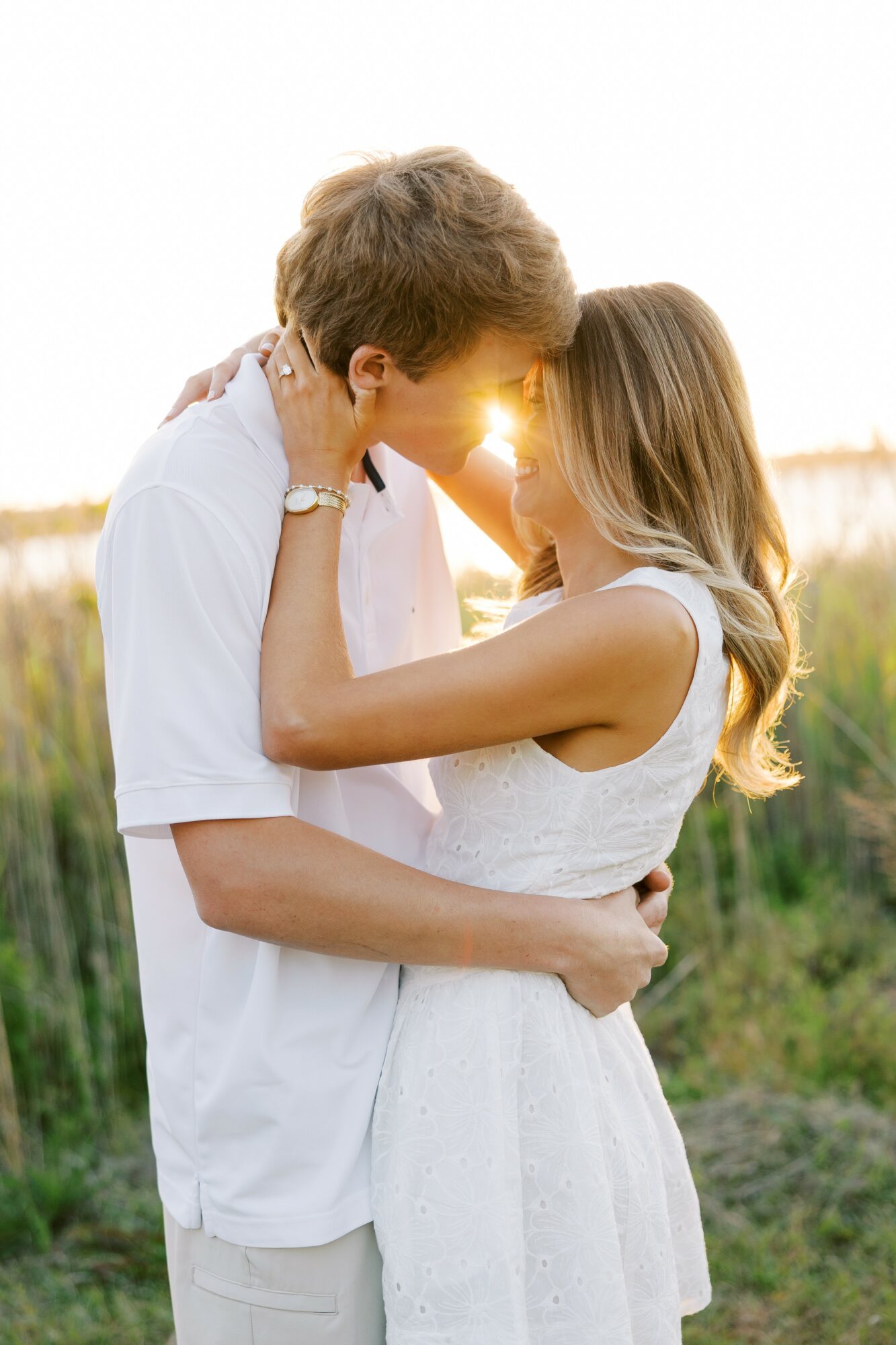 A couple stands close together outdoors, touching foreheads with sunlight behind them, in a natural setting with greenery.