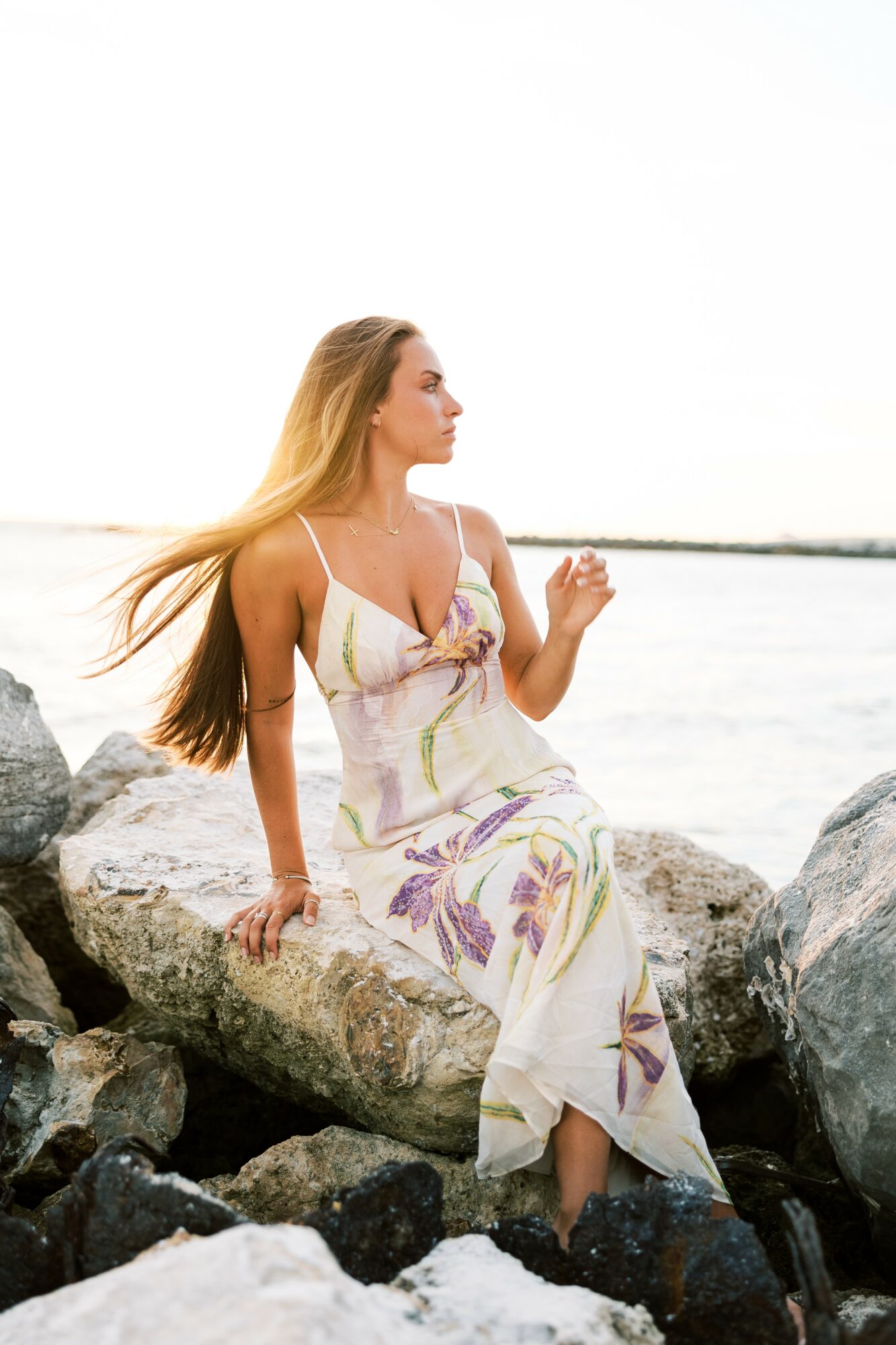 Woman with long hair sitting on rocks by water, wearing a light-colored dress with purple and green patterns, looking to the side.