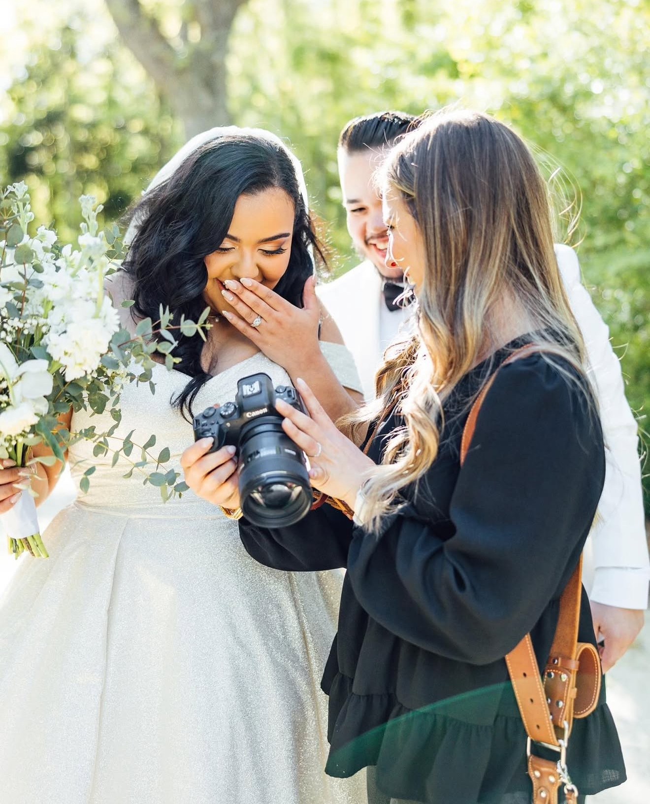 Three women outdoors, one holding a camera, smiling and looking at a photo, surrounded by greenery and flowers.