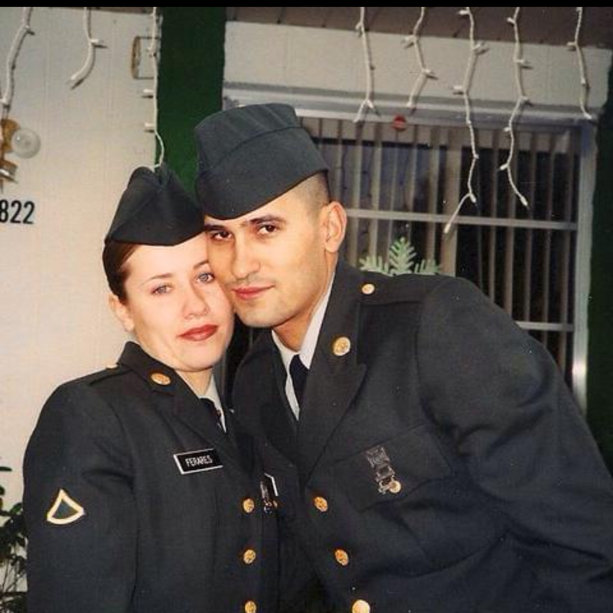 Two uniformed individuals, a woman and a man, standing close together and smiling, with holiday lights in the background.