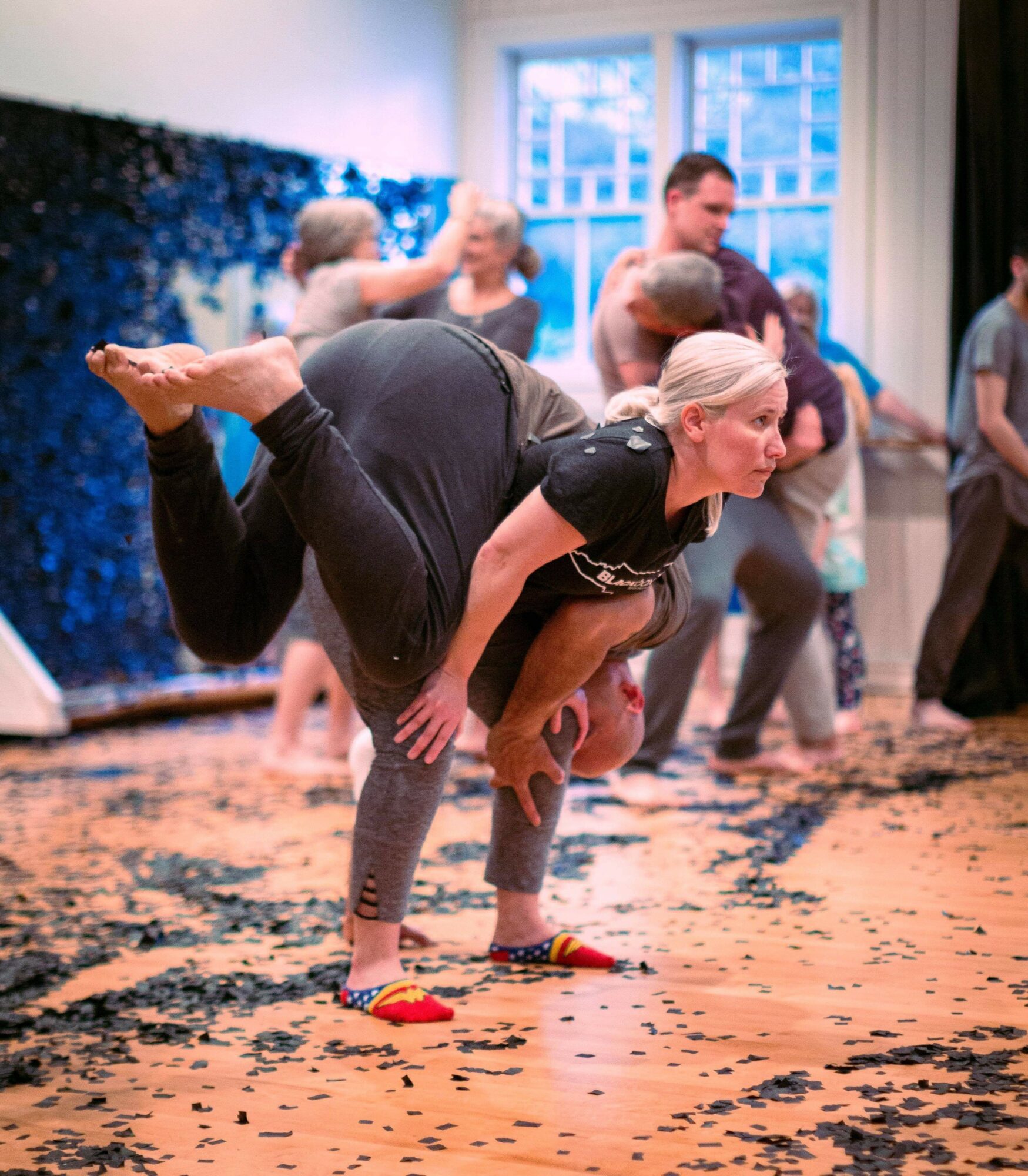 Woman balancing on one leg with the other leg extended backward, in a room with people in the background.