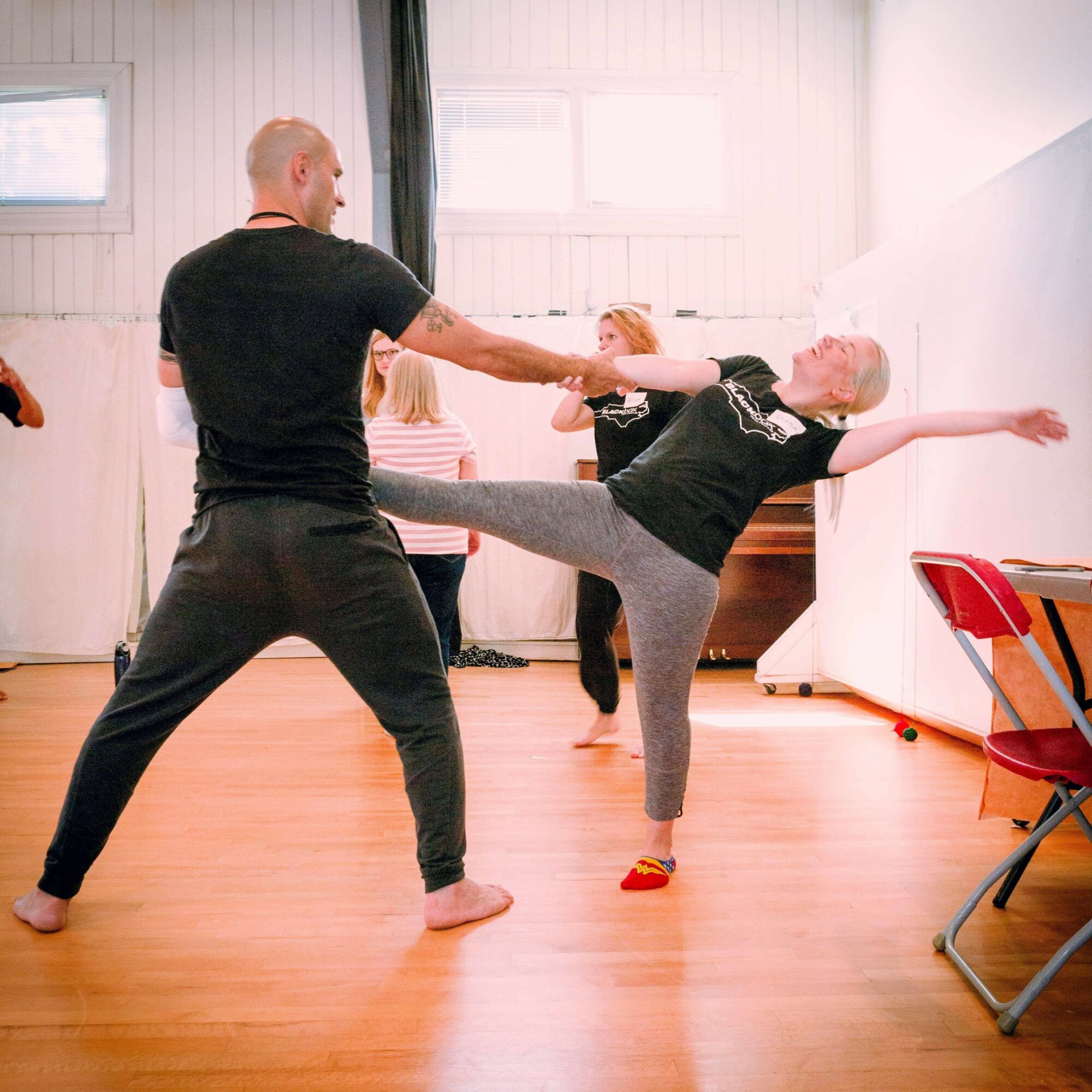 Adult and child practicing dance or exercise in a studio, child extending leg and arm, adult holding child's hand, others in background.