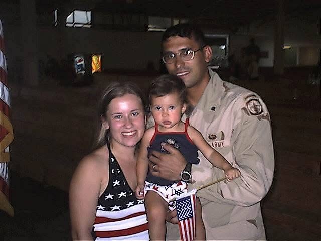 Three people, a woman, a man in uniform, and a young child, pose together indoors, smiling at the camera.