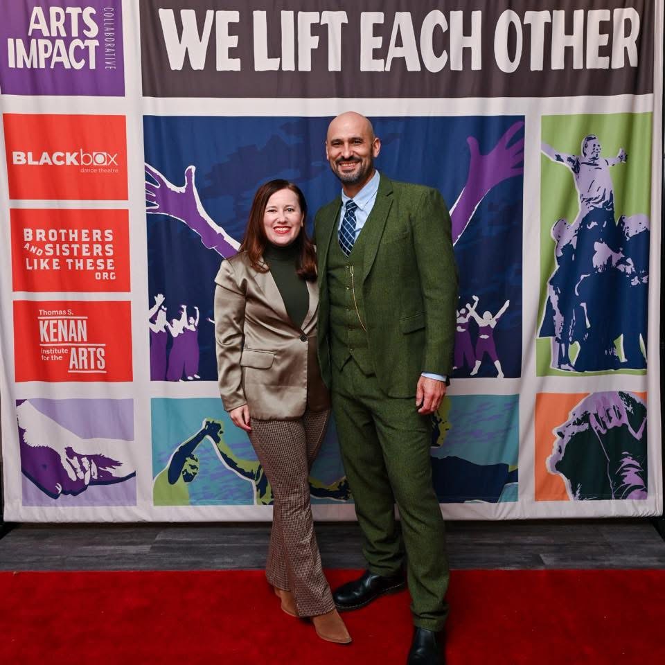 Two people, a woman and a man, stand on a red carpet in front of a colorful backdrop with text and graphics, smiling at the camera.