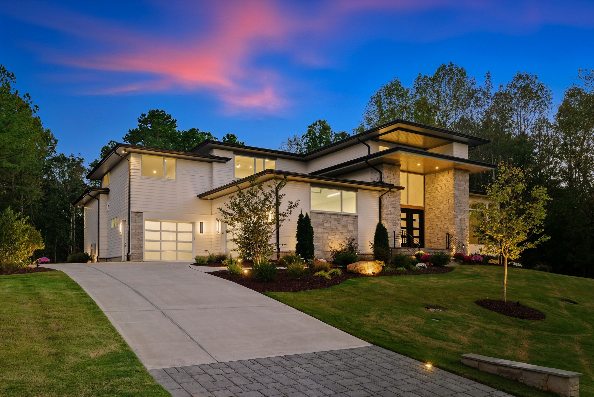 Modern house with large windows, driveway, and landscaped yard during sunset, surrounded by trees.