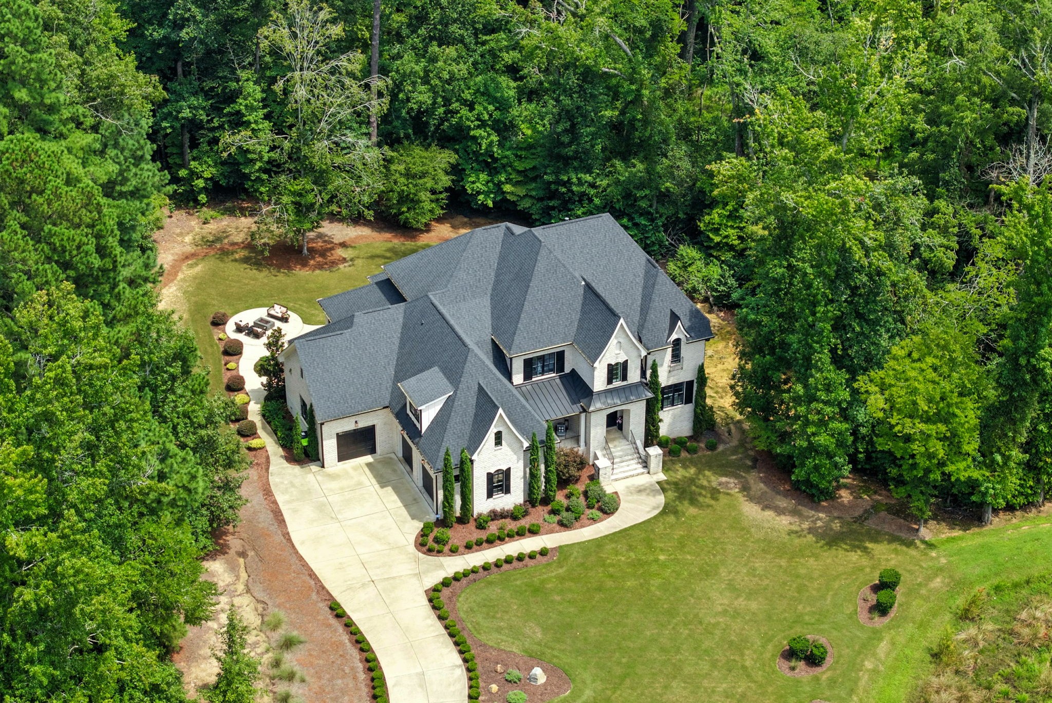A large house with a gray roof surrounded by trees and a lawn, with a driveway and parked cars nearby.
