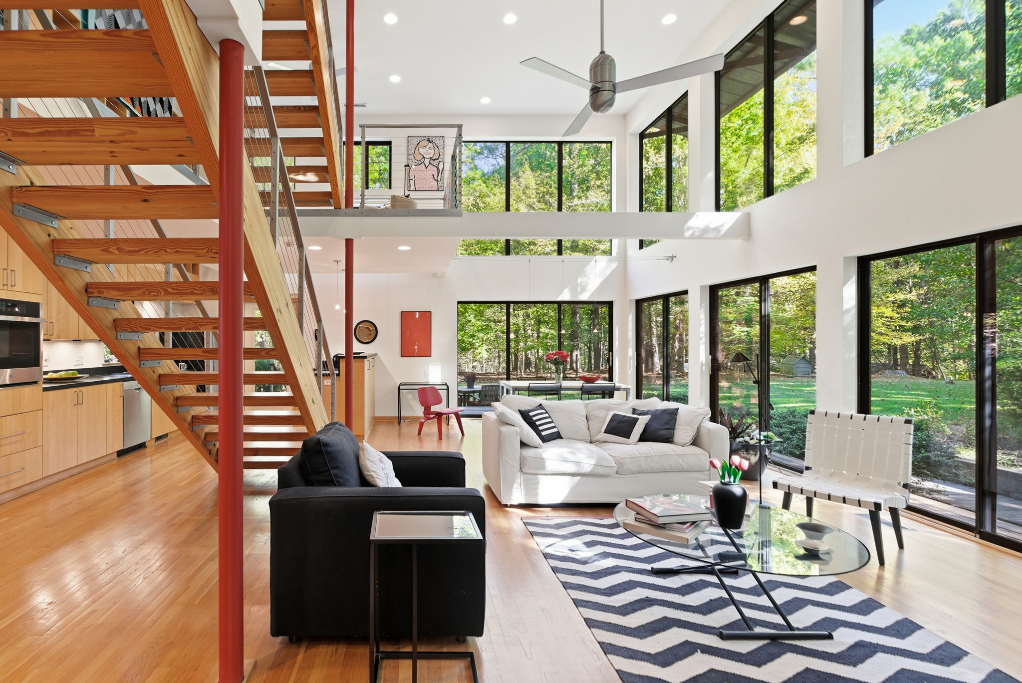 Bright living room with large windows, white and black sofas, wooden staircase, and a chevron-patterned rug.