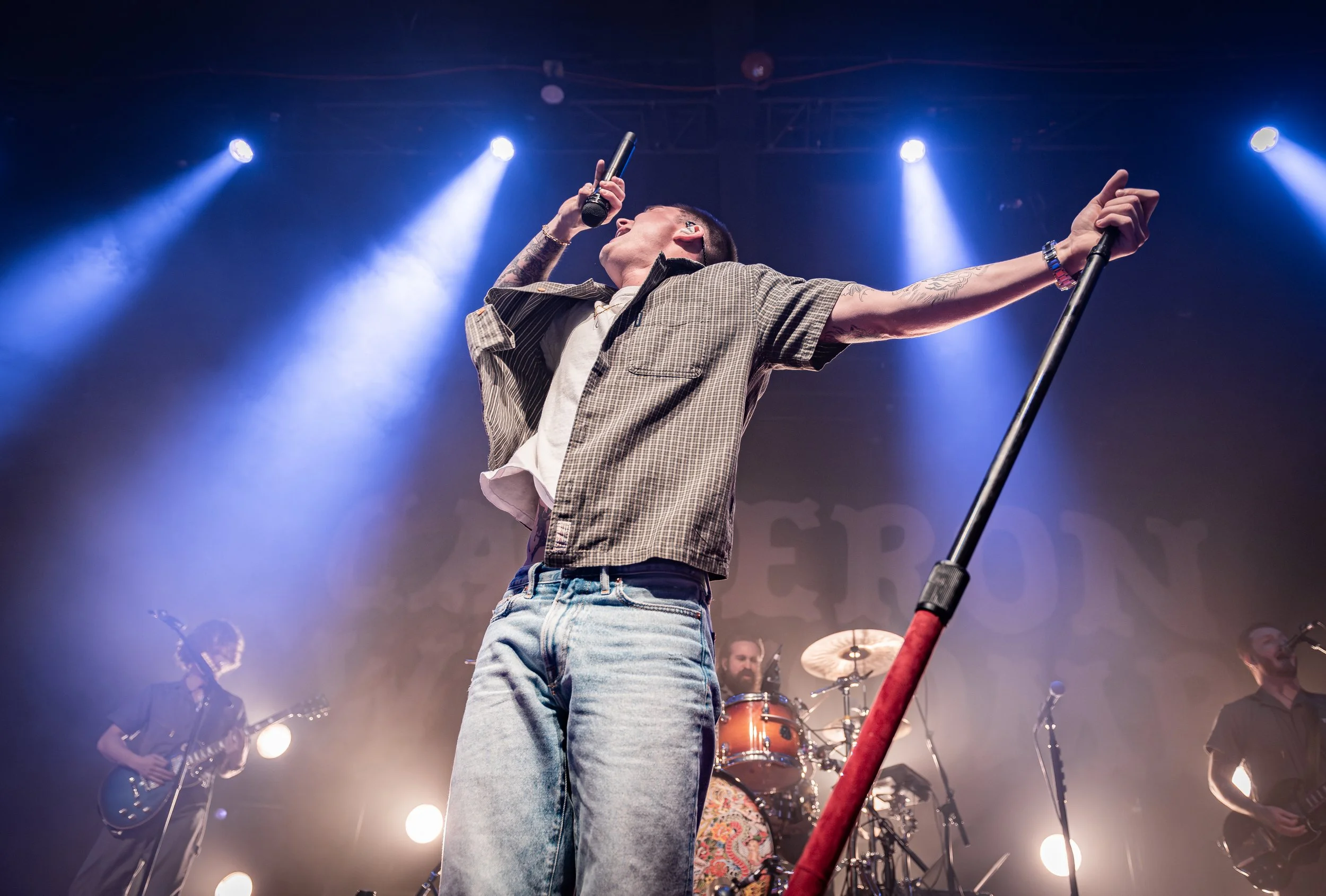 Performer singing on stage with microphone, holding a pole, with band members and stage lights in background.
