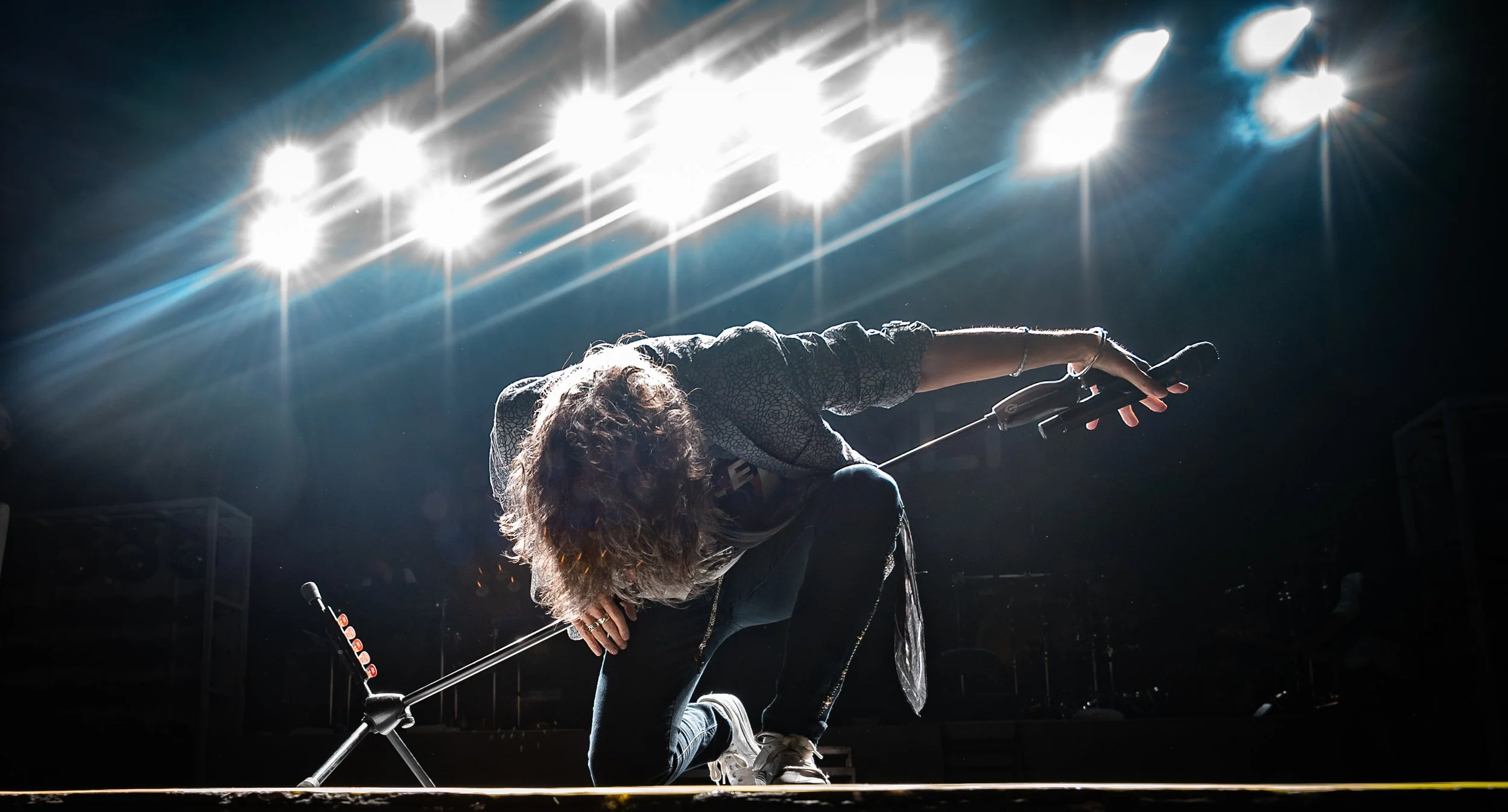 Person with curly hair crouching on stage with bright lights overhead, stage equipment visible in background.
