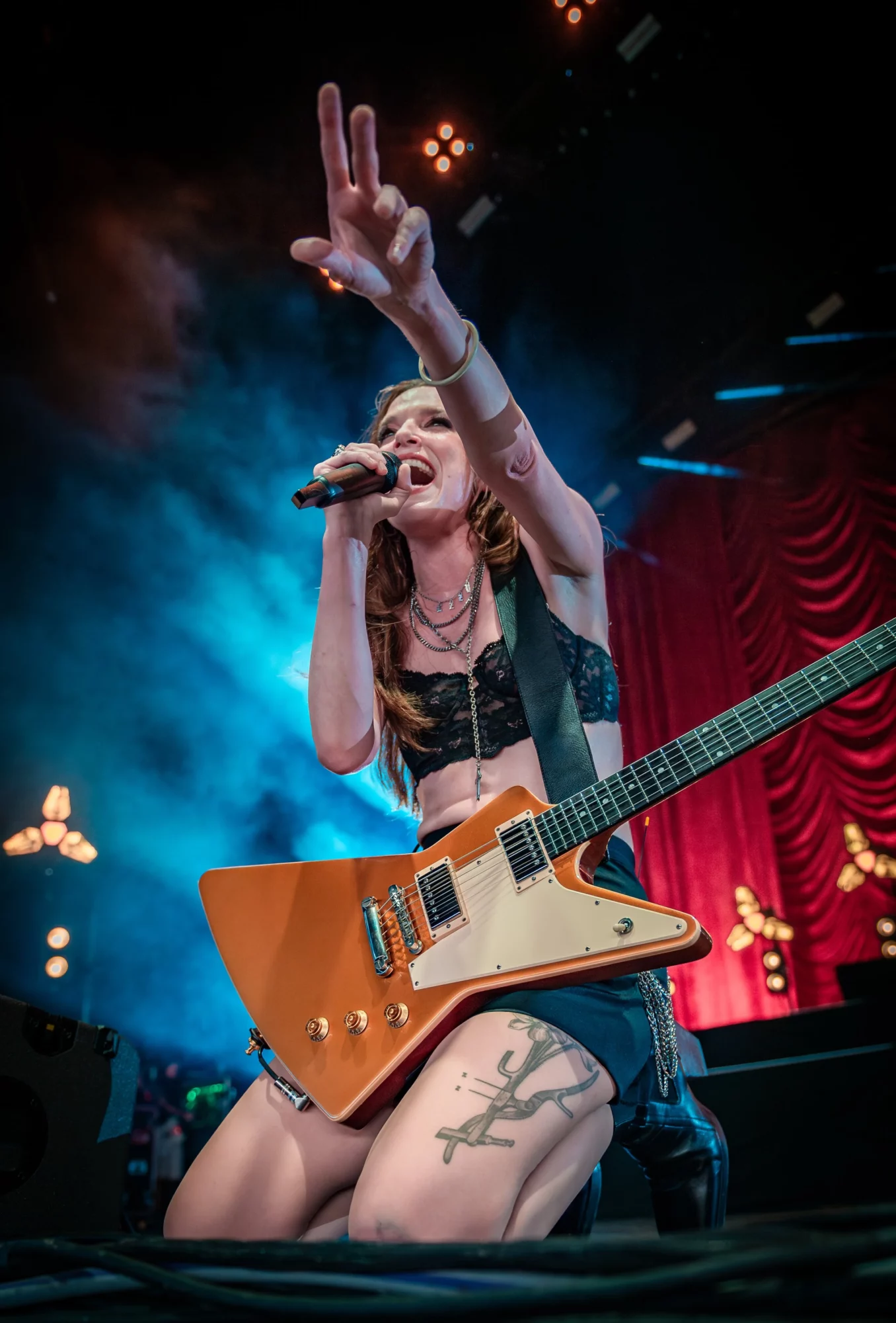 Female musician kneeling on stage with a guitar, singing into a microphone, with stage lights and red curtain background.