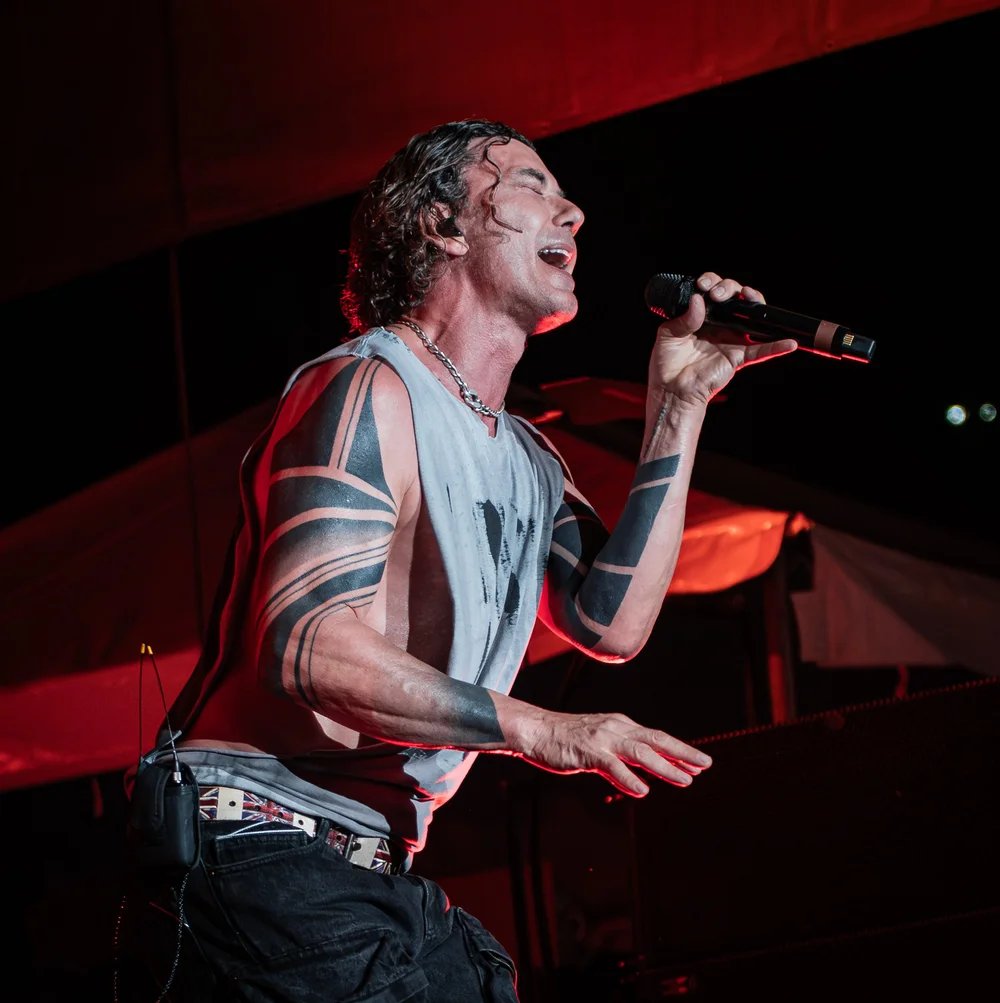 Person singing into a microphone on stage with red lighting, wearing a sleeveless top and showing tattoos on arms.