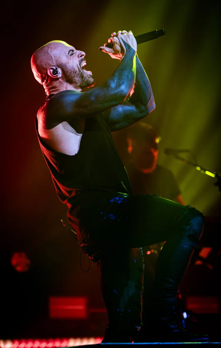 Male singer with a shaved head and tattoos performs passionately on stage, holding a microphone, with colorful stage lighting.