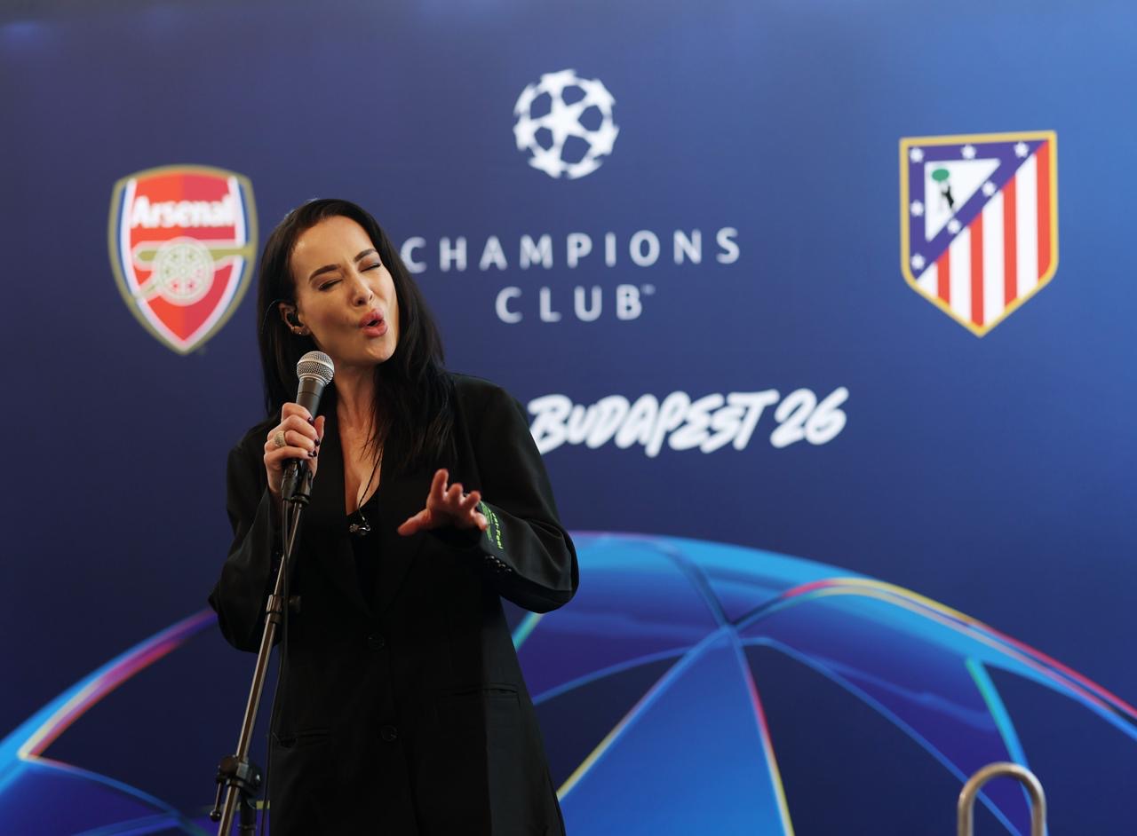 Woman speaking into a microphone in front of a UEFA Champions League backdrop with Arsenal and Atlético Madrid logos.