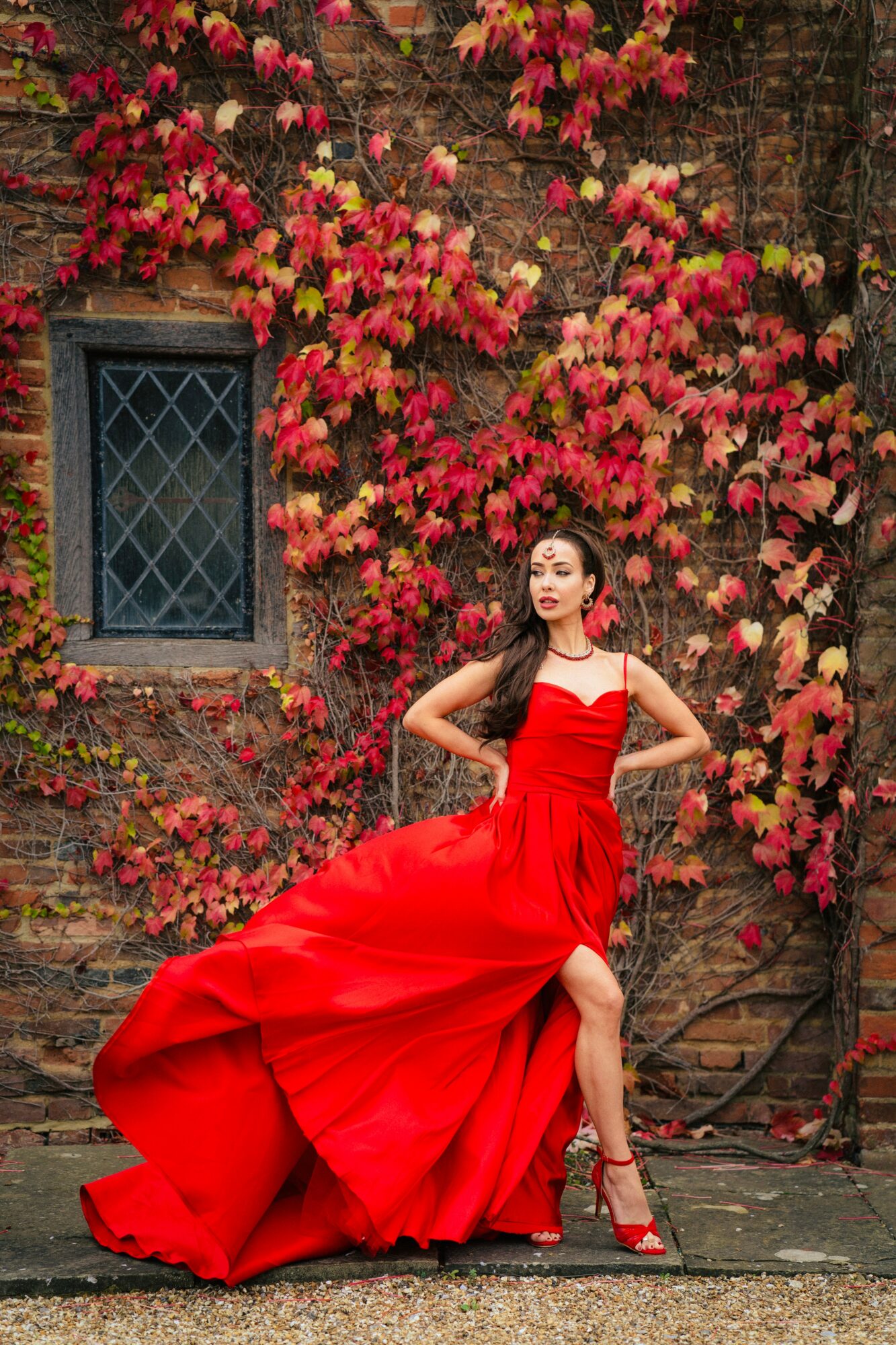 Woman in a red gown standing in front of a brick wall with colorful climbing plants, posing confidently.