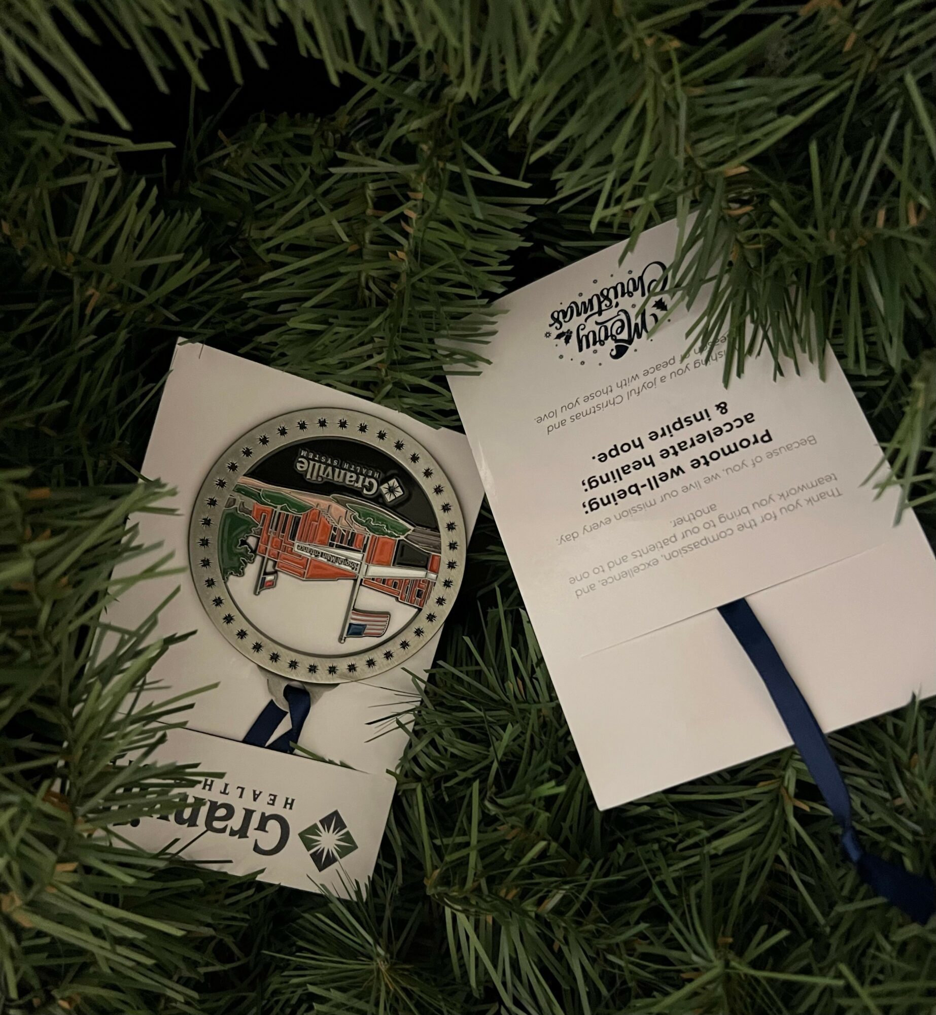 Two cards placed among green pine branches, one showing a circular logo with a building and the other with text and a ribbon.