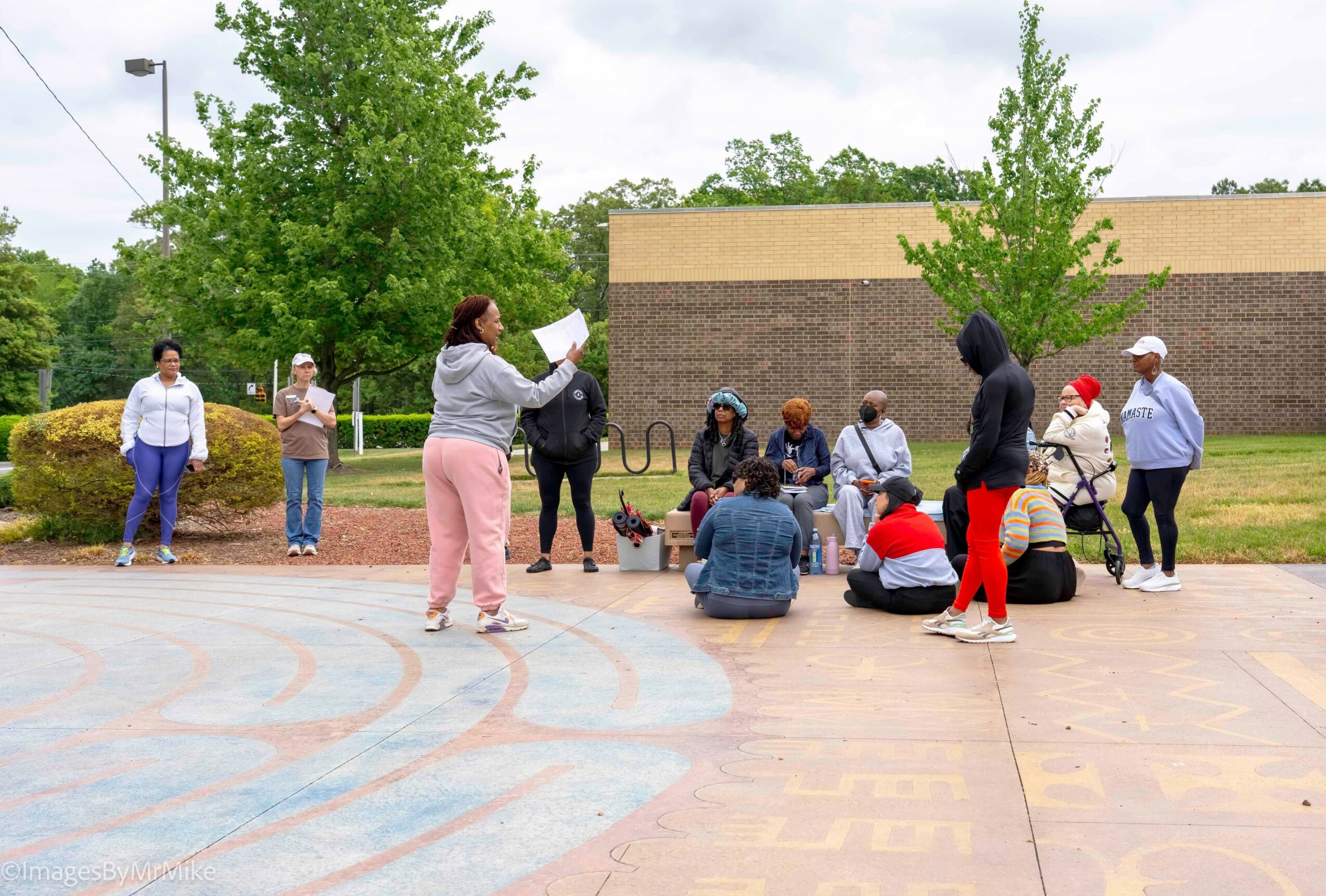 Group of people sitting and standing outdoors, some listening to a woman reading a paper, trees and a building in background.