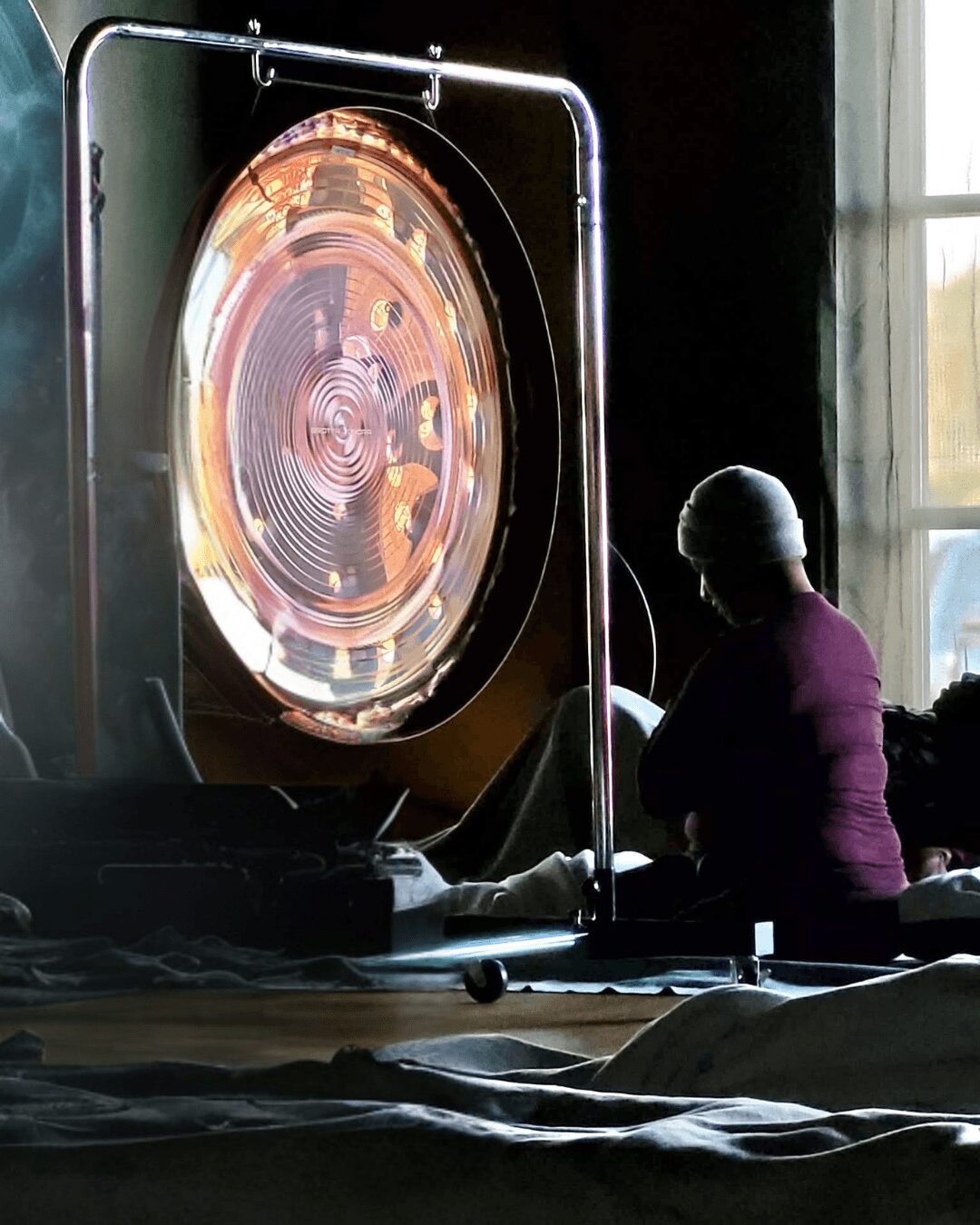 Person sitting near a large, circular, illuminated device with concentric rings, inside a room with a window, facing away.
