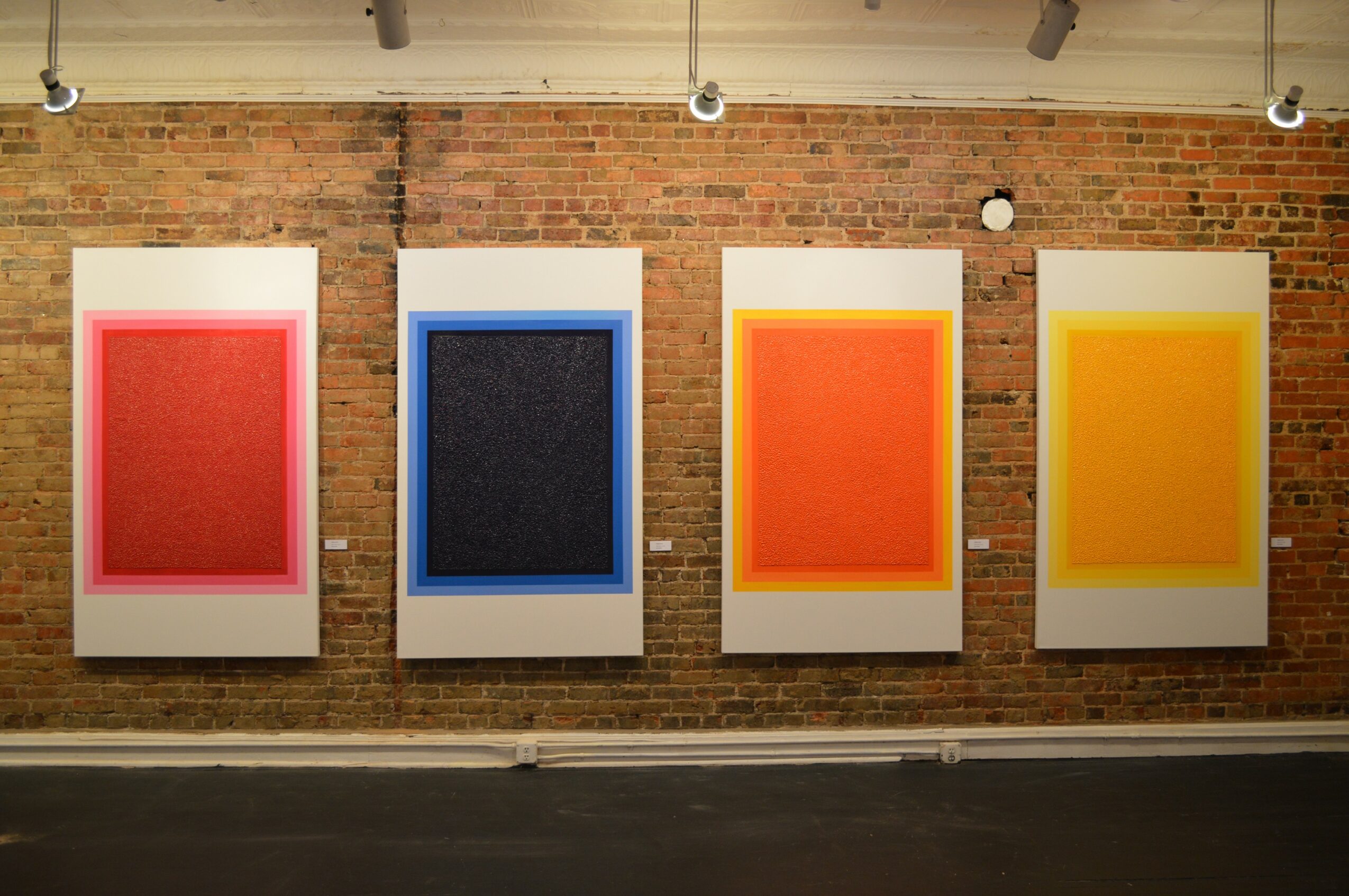 Four framed abstract art pieces with colorful textured backgrounds hang on a brick wall in a gallery.