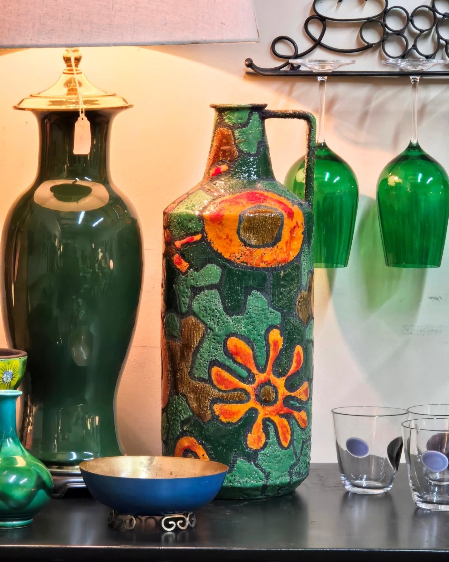 Decorative ceramic vase with colorful floral patterns, flanked by green glass bottles and other glassware, against a pink wall.